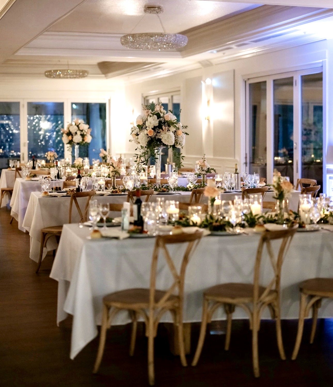 Elegant banquet hall decorated for a wedding or special event with long tables, candle centerpieces, floral arrangements, and string lights, lit warmly at night.
