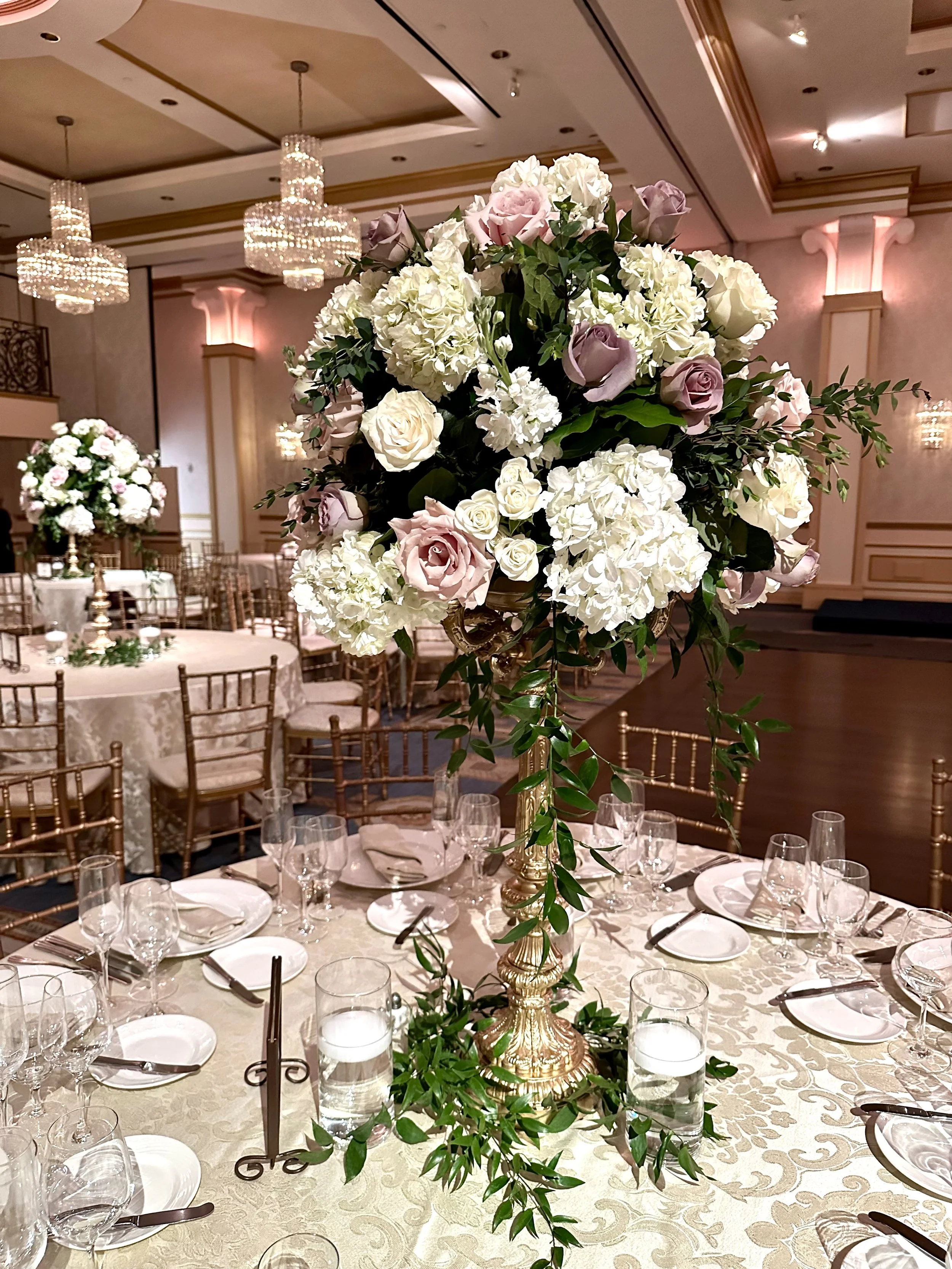 A large floral centerpiece with white hydrangeas, roses, and greenery on a table at a formal event in a decorated banquet hall with chandeliers and elegant lighting.