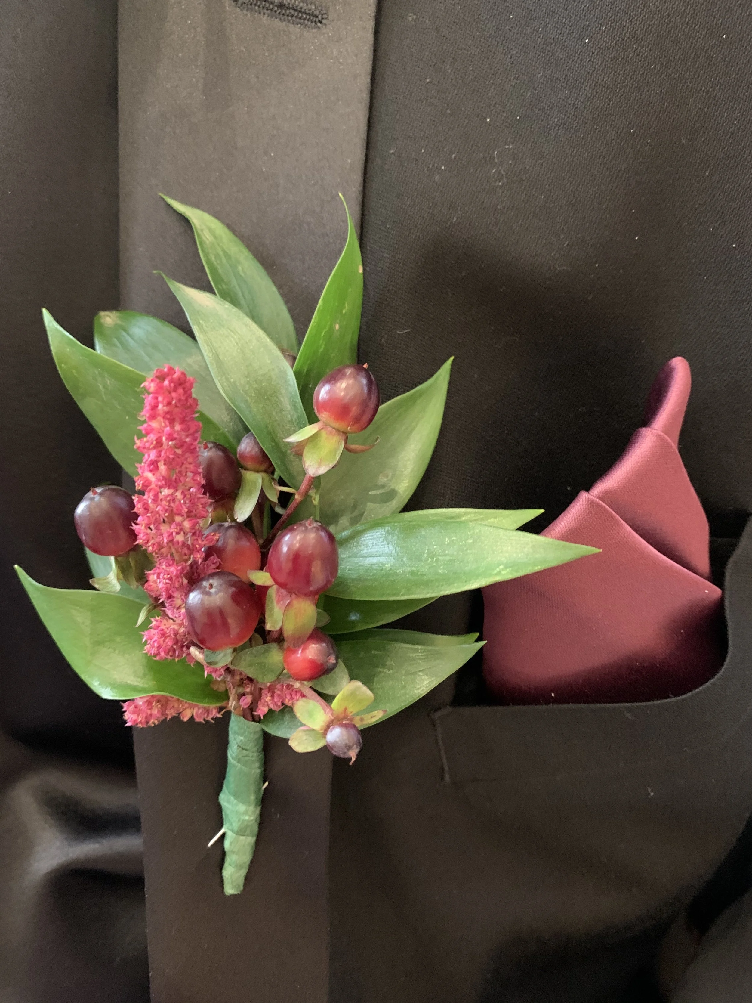 A boutonniere with pink and red flowers and green leaves resting on a black fabric surface with a burgundy napkin.