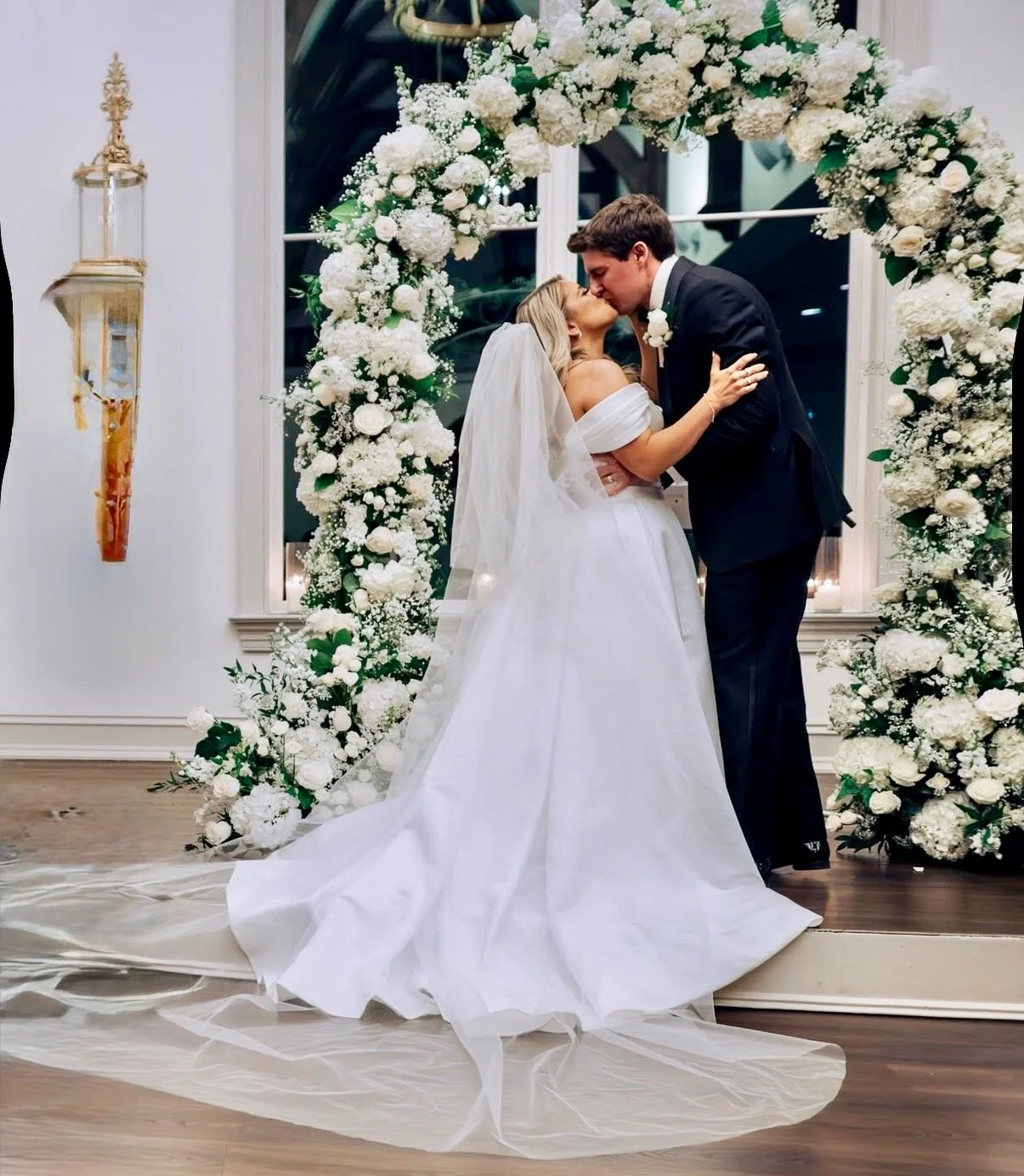 A bride and groom sharing a kiss during their wedding ceremony, standing under a white floral arch with white flowers and greenery inside a decorated room.