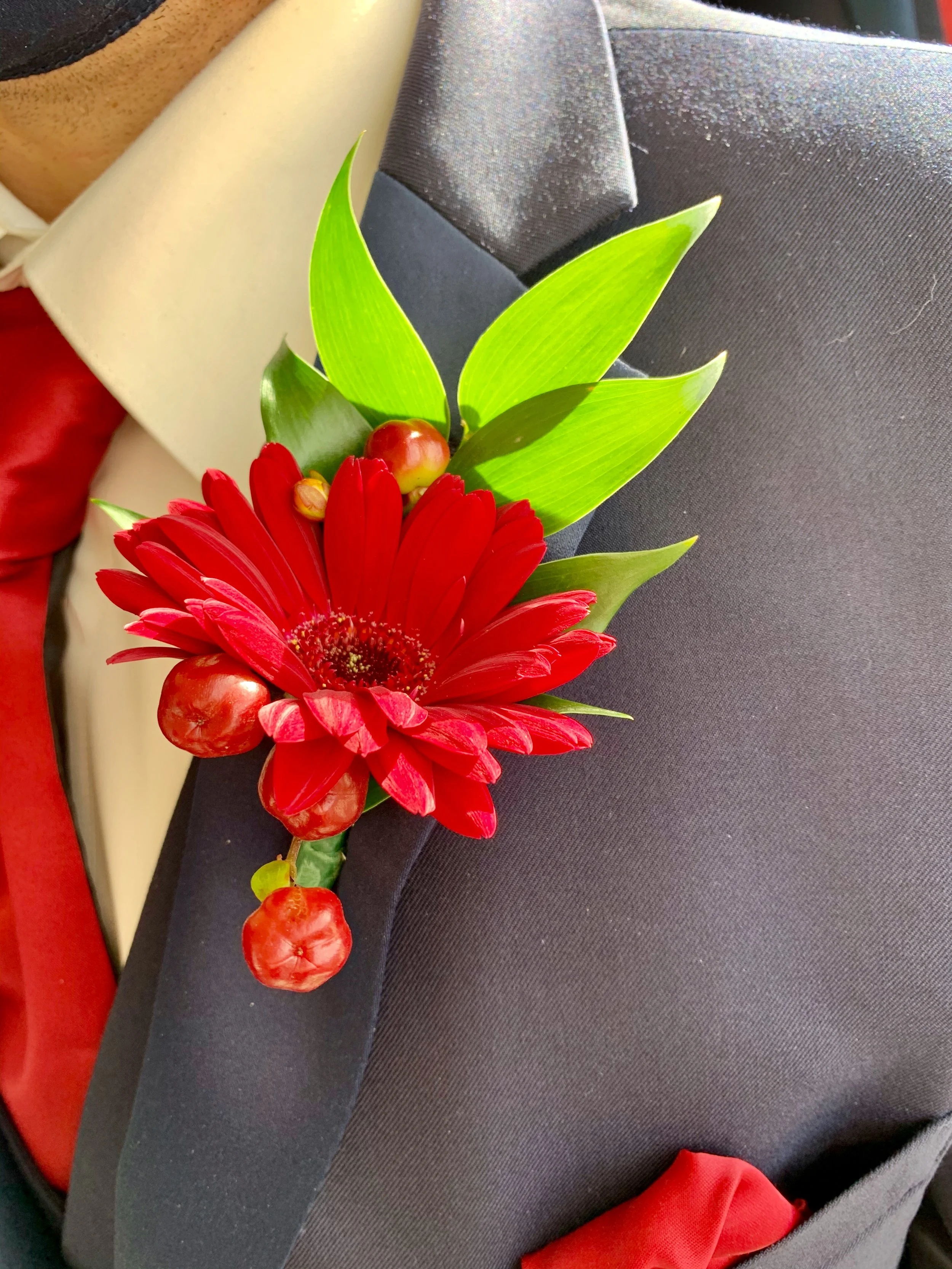 Close-up of a boutonniere with a red flower, green leaves, and red berries, attached to a dark suit jacket.