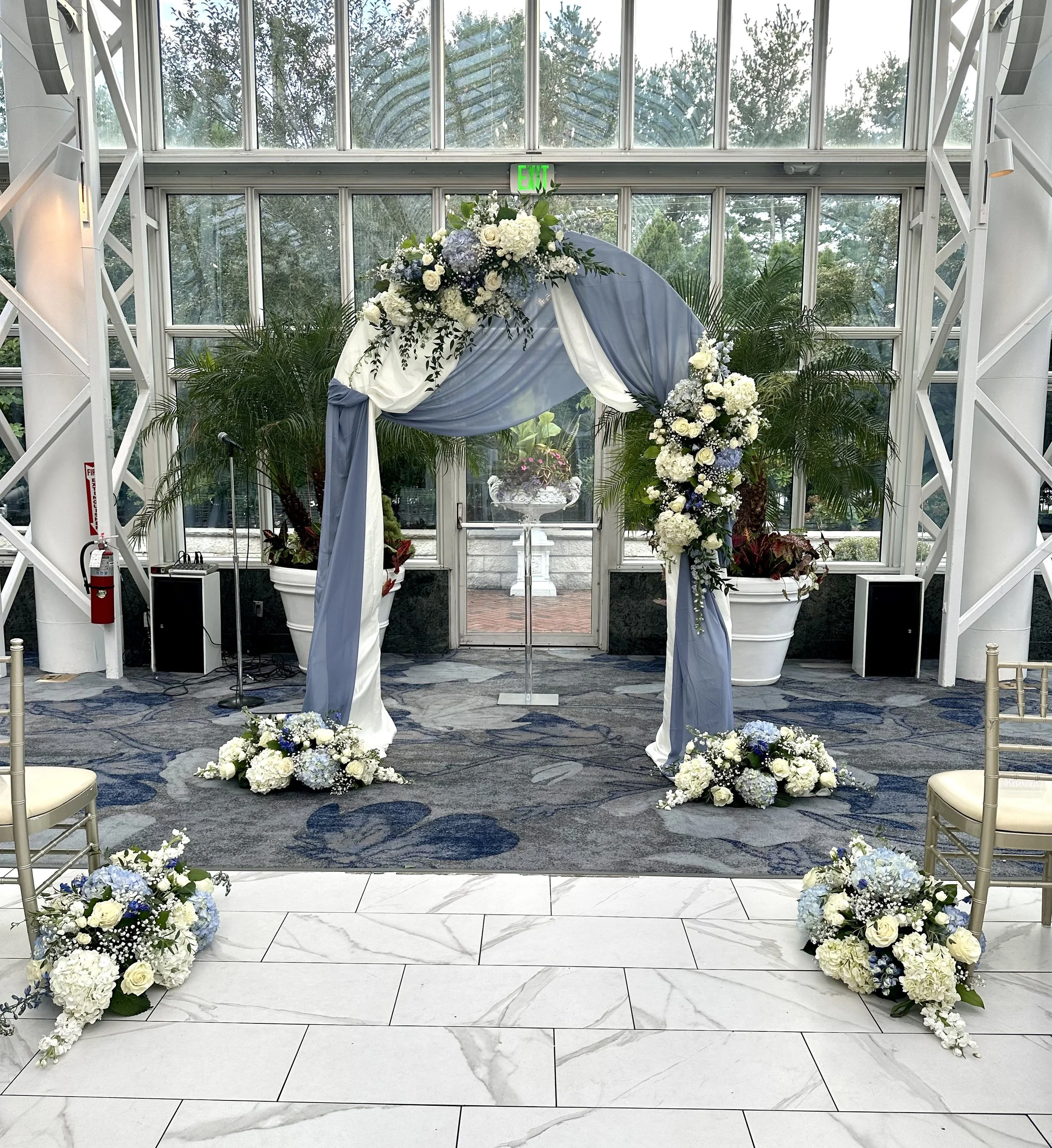 Indoor wedding ceremony setup with flower arrangements, draped fabric, and an arch, inside a glass-enclosed space with plants and trees outside.