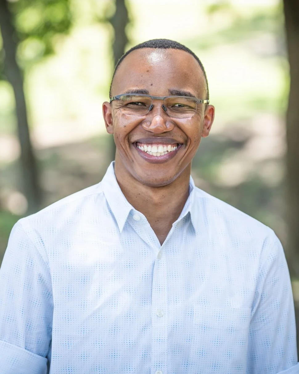 A smiling man wearing glasses and a white dress shirt, outdoors with blurred greenery in the background.