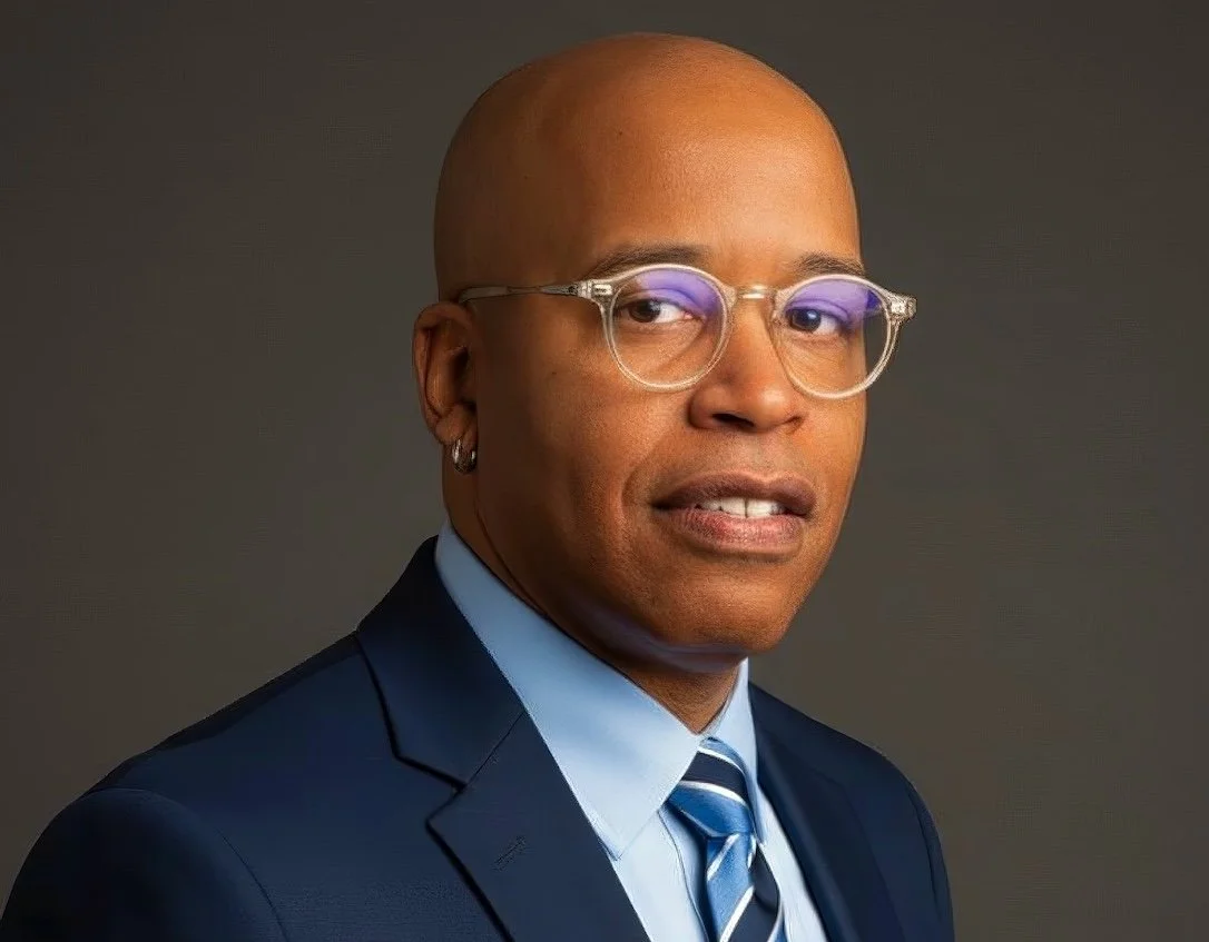 Portrait of a bald African American man with clear glasses wearing a navy suit, light blue shirt, and striped tie against a dark background.