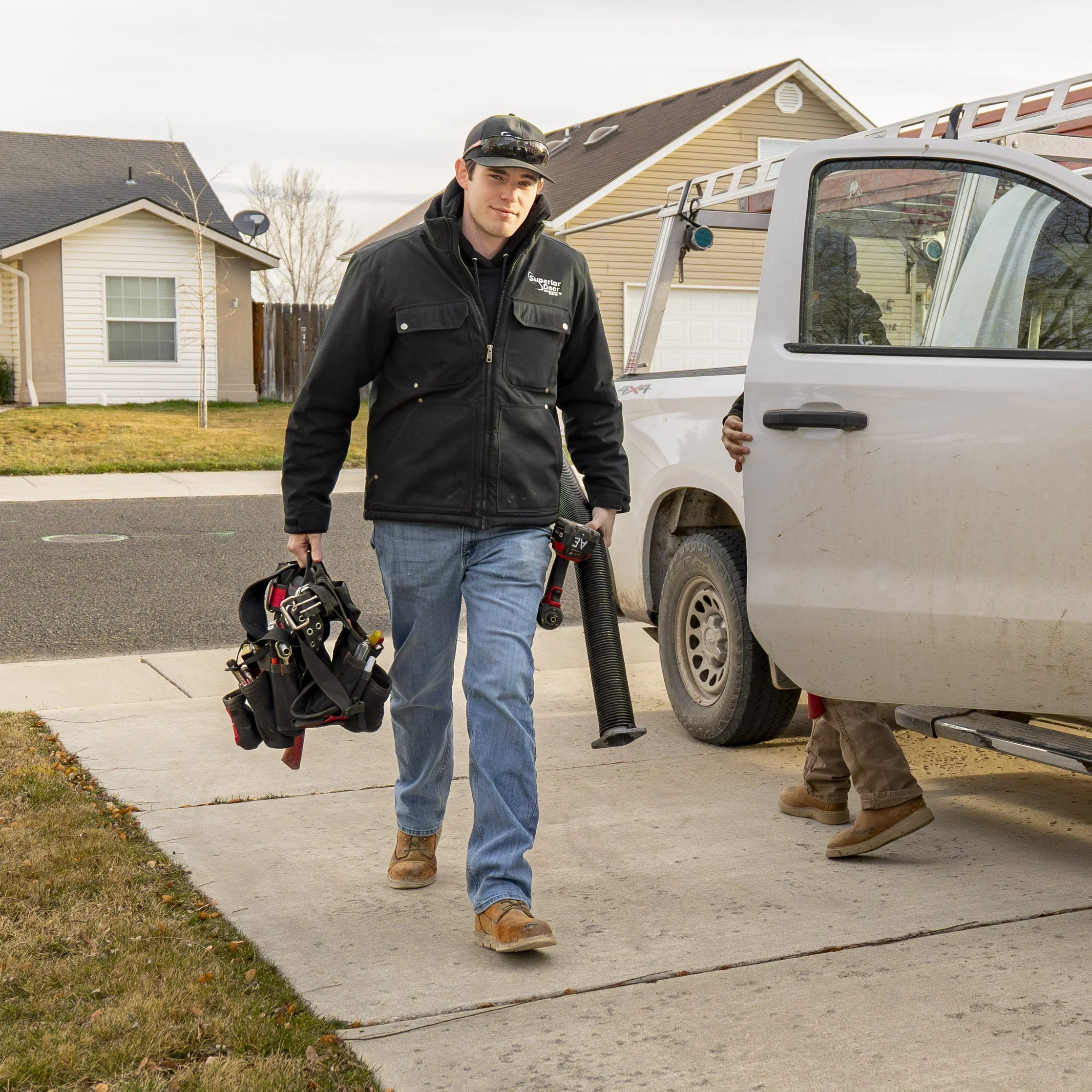 A man in work clothes and boots walking on a driveway, carrying a toolbox and using a vacuum hose, with houses in the background.