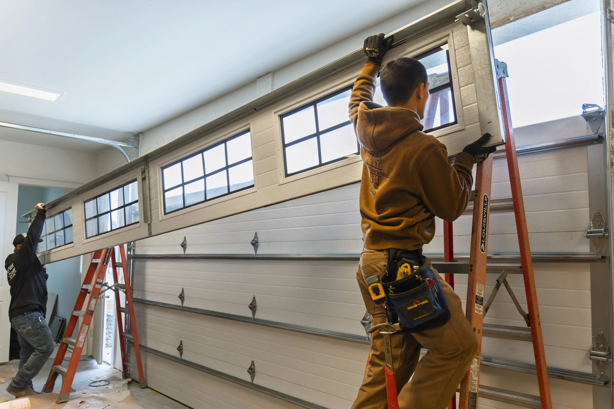 Construction workers installing a garage door, one worker on an orange ladder and another on the ground, inside a garage with rectangular window panels.