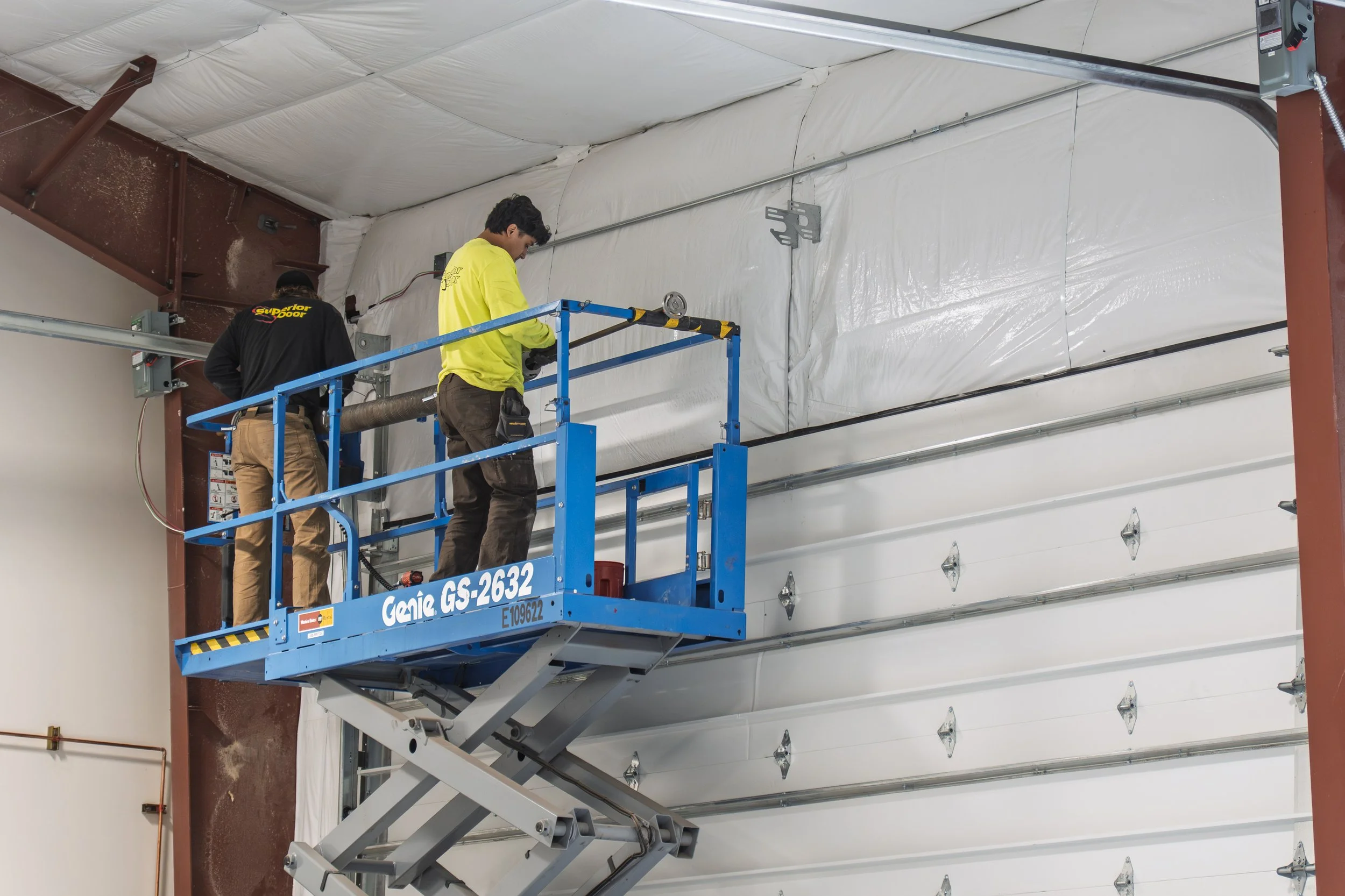 Two workers are standing on a blue scissor lift in a large indoor space with a high ceiling and a white wrapping on the walls. One worker, wearing a yellow shirt, is using a power tool, while the other, wearing a black jacket with yellow text, is facing away from the camera.