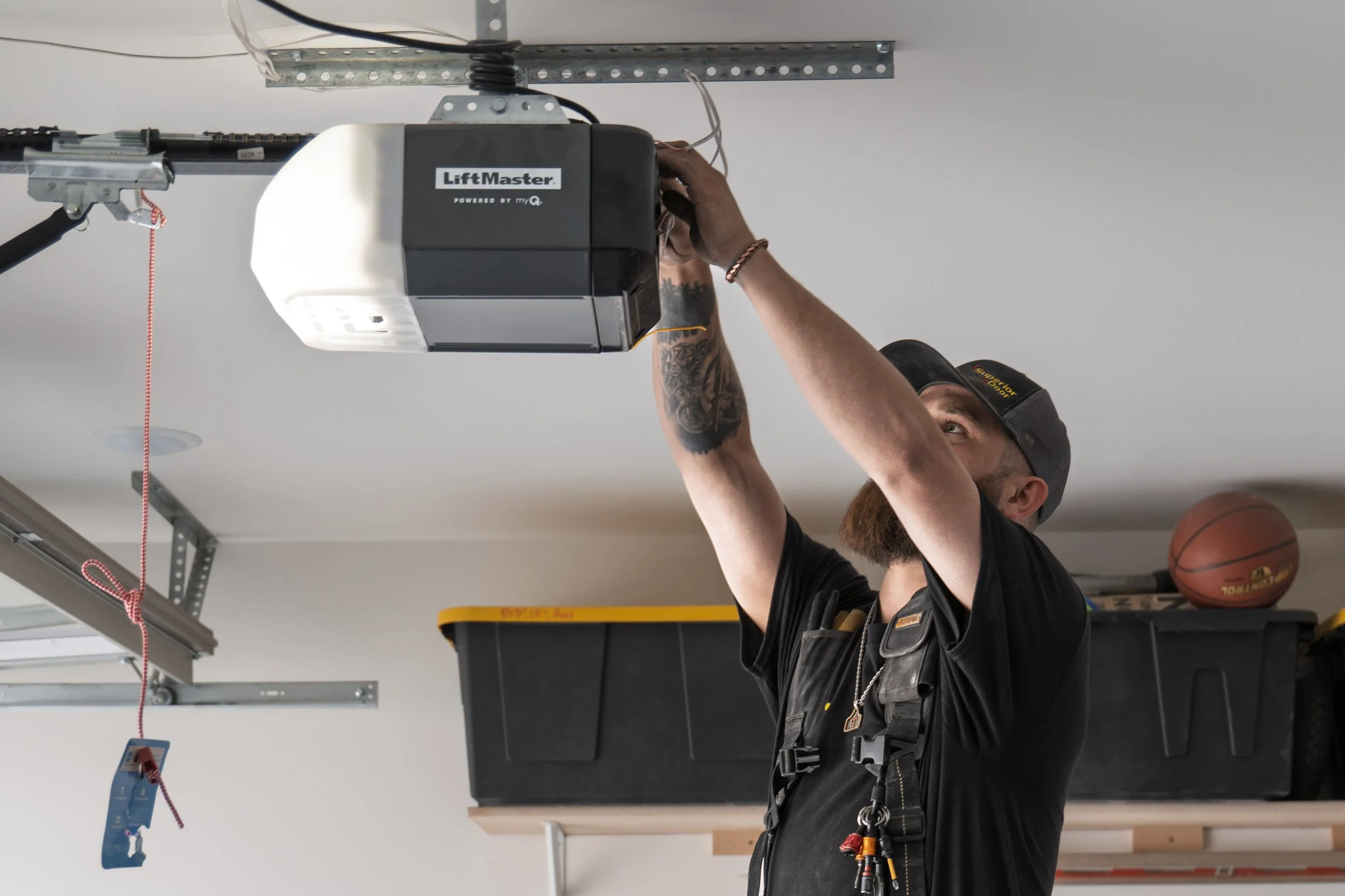 A person in a garage installing or repairing a LiftMaster garage door opener mounted on the ceiling. The individual is wearing a black cap and work vest, and is handling wires near the opener.