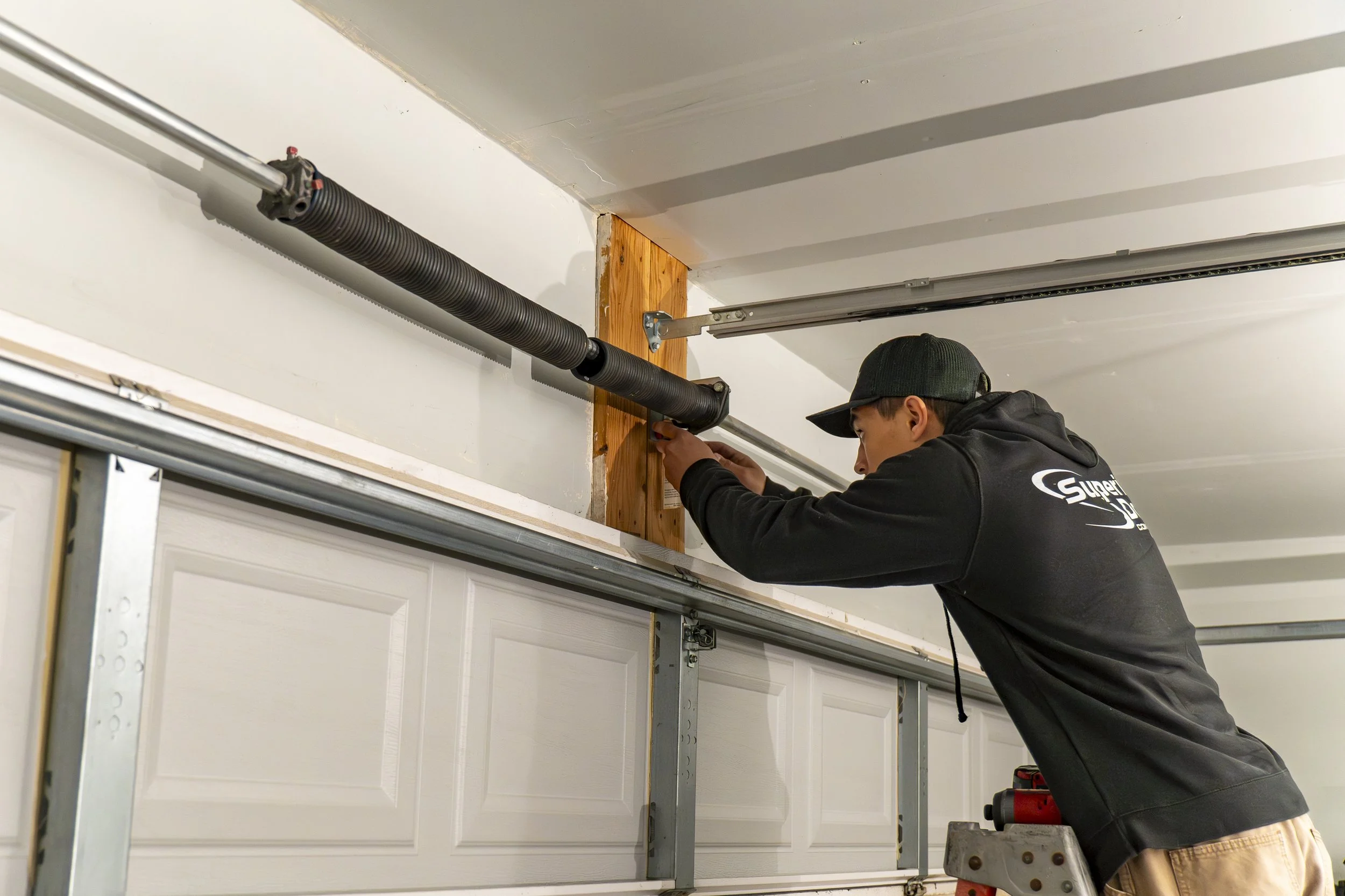 A person wearing a black hoodie and cap is working on installing a garage door opener track. They are using a power drill and are positioned on a step ladder in a garage, with a wood support beam and the garage door track visible.