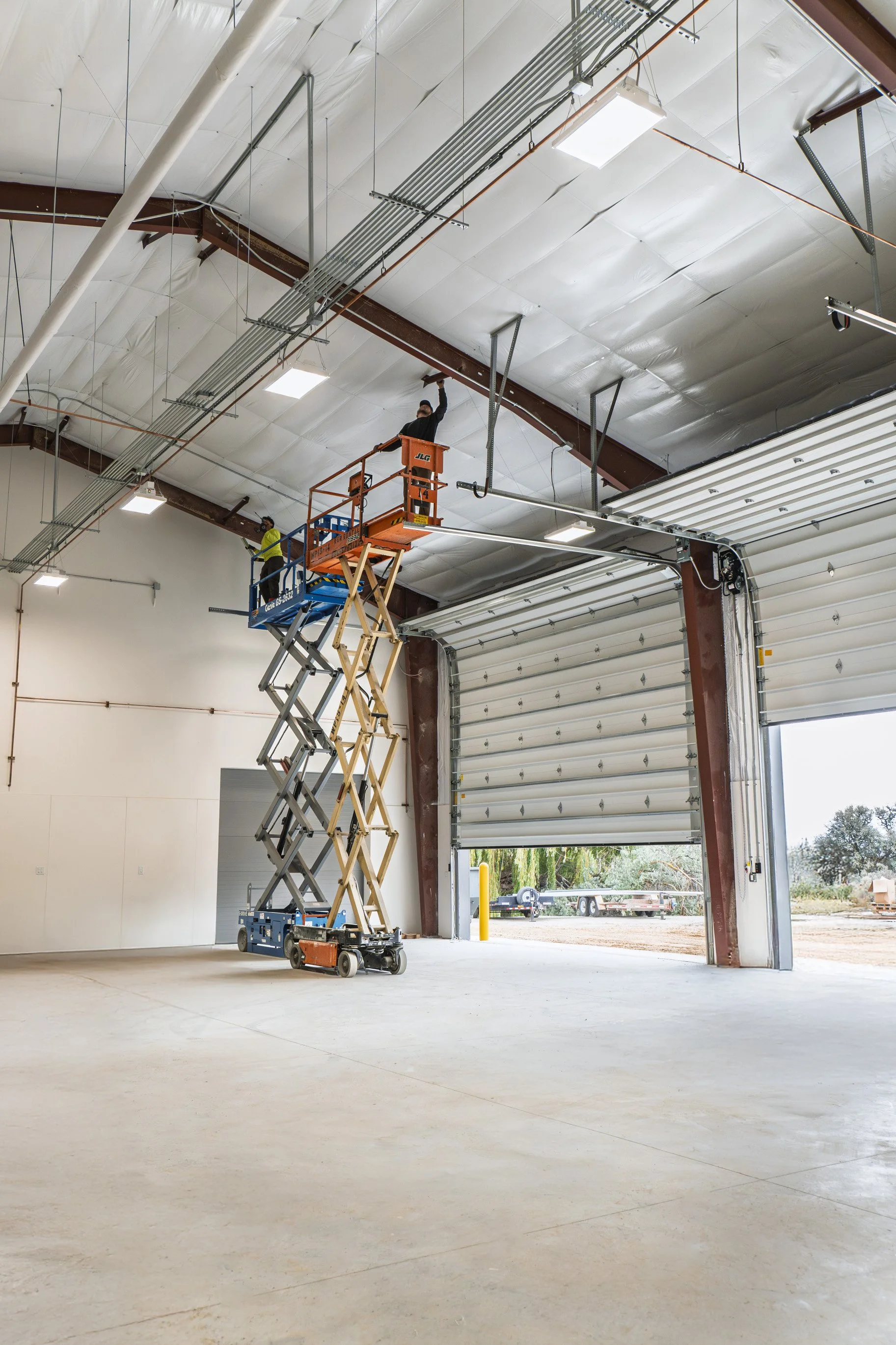 Inside a warehouse with an open garage door, a worker on a scissor lift is installing or repairing ceiling fixtures, while another worker stands nearby. The warehouse has a steel structure and the floor is concrete.