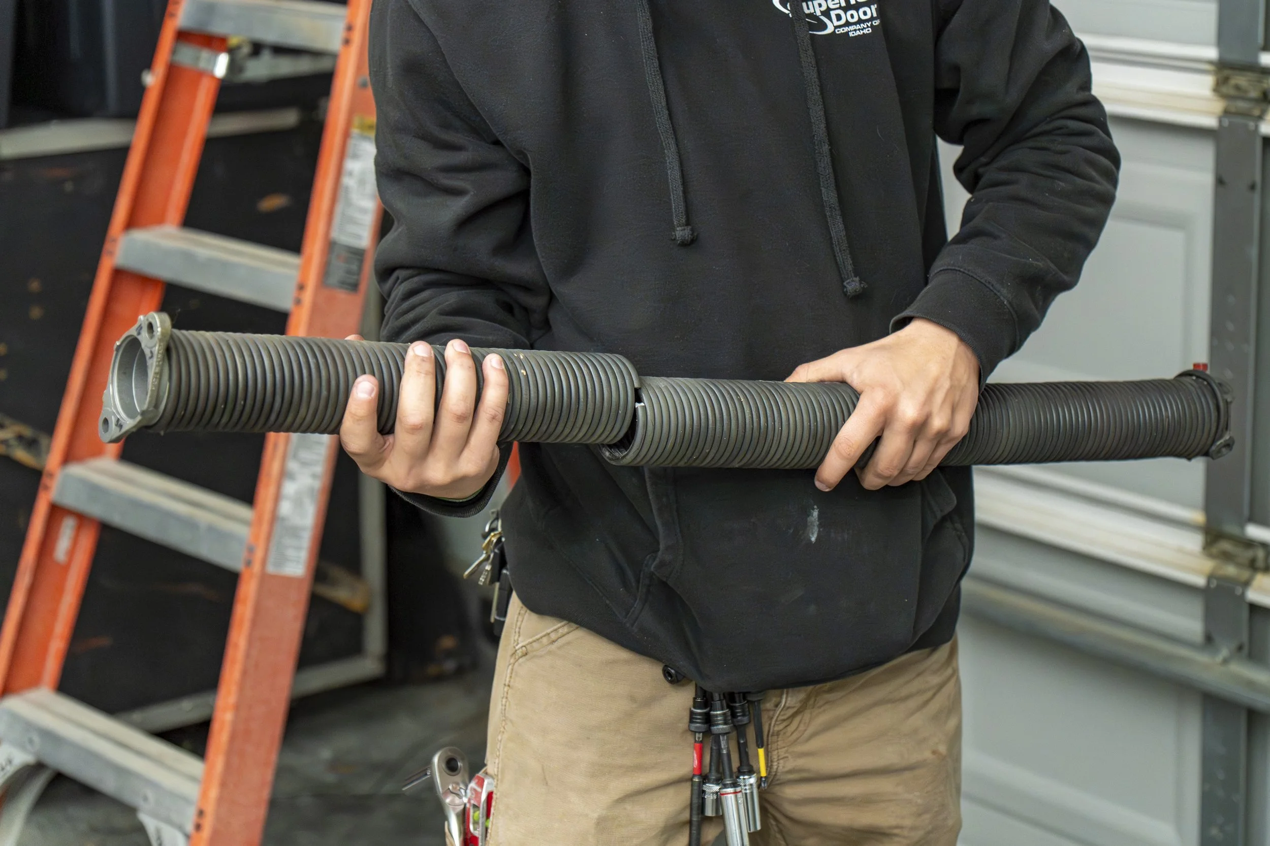 A person in a black hoodie holding a ribbed, cylindrical metal pipe in an industrial or workshop setting.