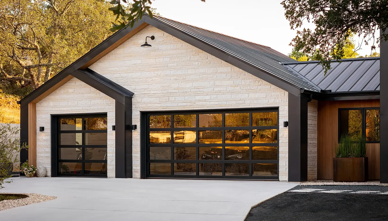 Modern house with white brick exterior, black garage doors with glass panels, metal roof, and wooden accents, surrounded by trees and a concrete driveway.