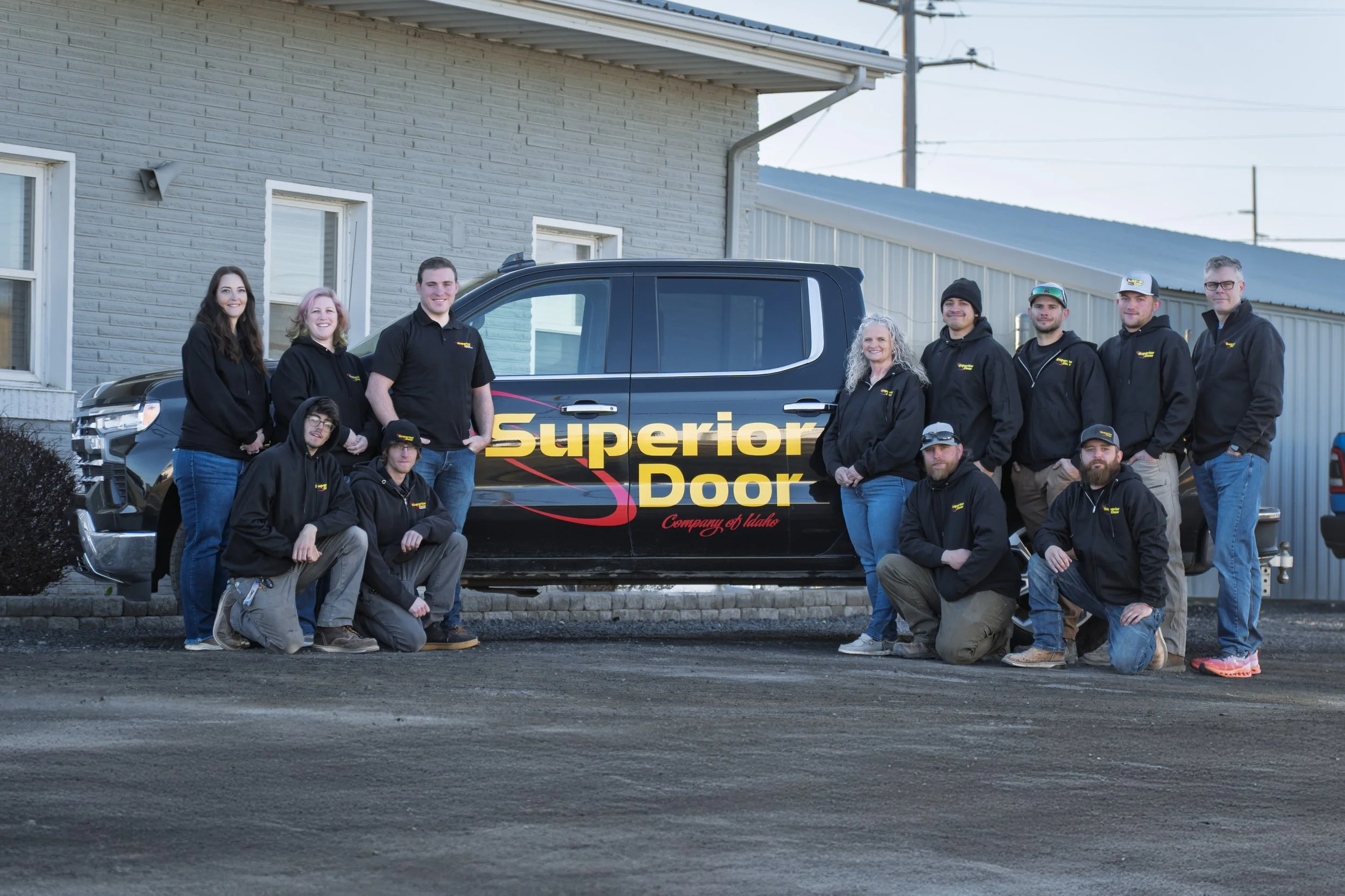 Group of nine people standing and kneeling in front of a company vehicle with 'Superior Door' logo, in an outdoor setting next to a brick building with windows and a metal siding building in the background.