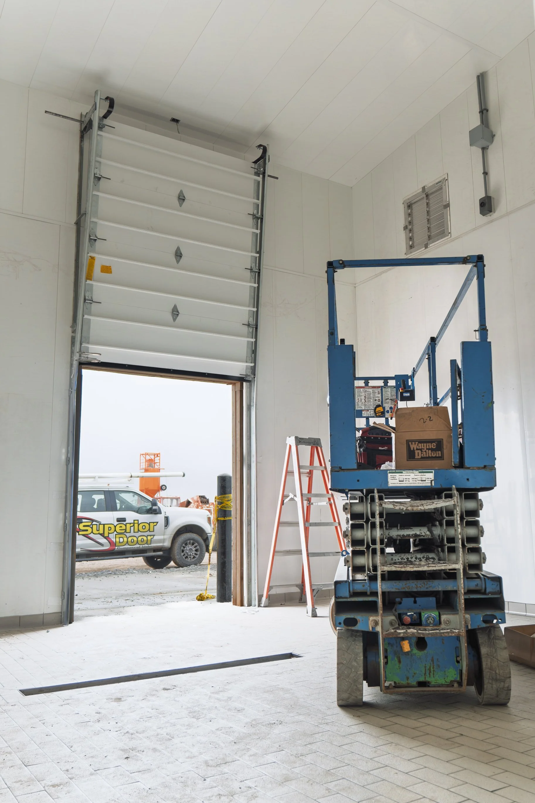 Construction site inside a building with a blue scissor lift, orange ladder, and workers outside near a white vehicle with 'Superior Door' logo, working on installing or repairing a garage door.