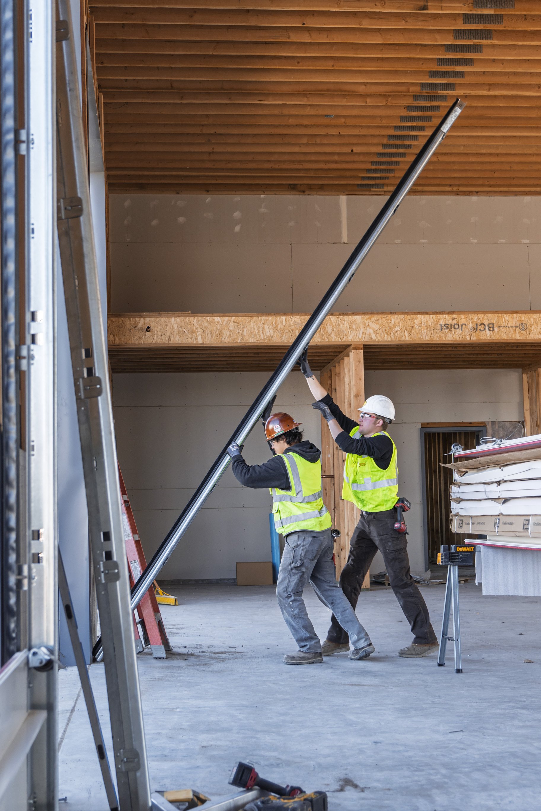 Two construction workers wearing hard hats, yellow safety vests, and gloves are working together inside a building under construction. One is holding a long metal pole, and the other is assisting. Construction materials and tools are scattered around, with wooden framing and partially finished walls visible in the background.