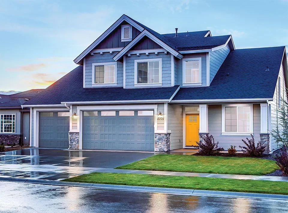 A modern two-story house with blue siding, stone accents, a yellow front door, a three-car garage, and a well-maintained lawn, with a wet driveway and overcast sky.