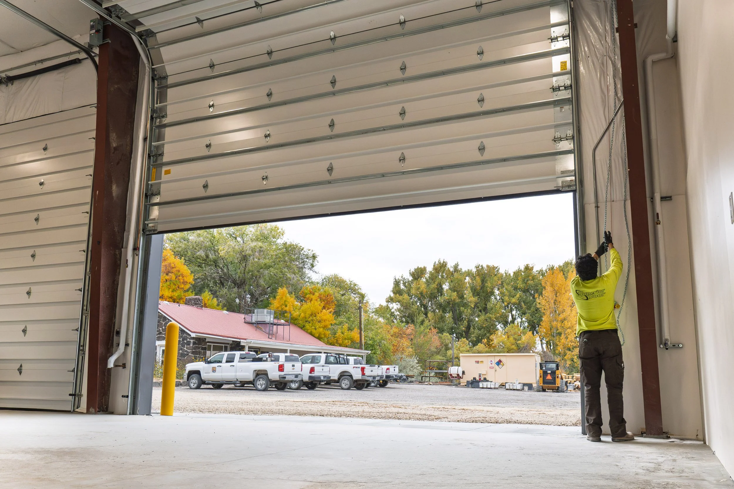 A person in a yellow shirt is operating a large industrial garage door, opening it to reveal a parking lot with several trucks and autumn-colored trees outside.