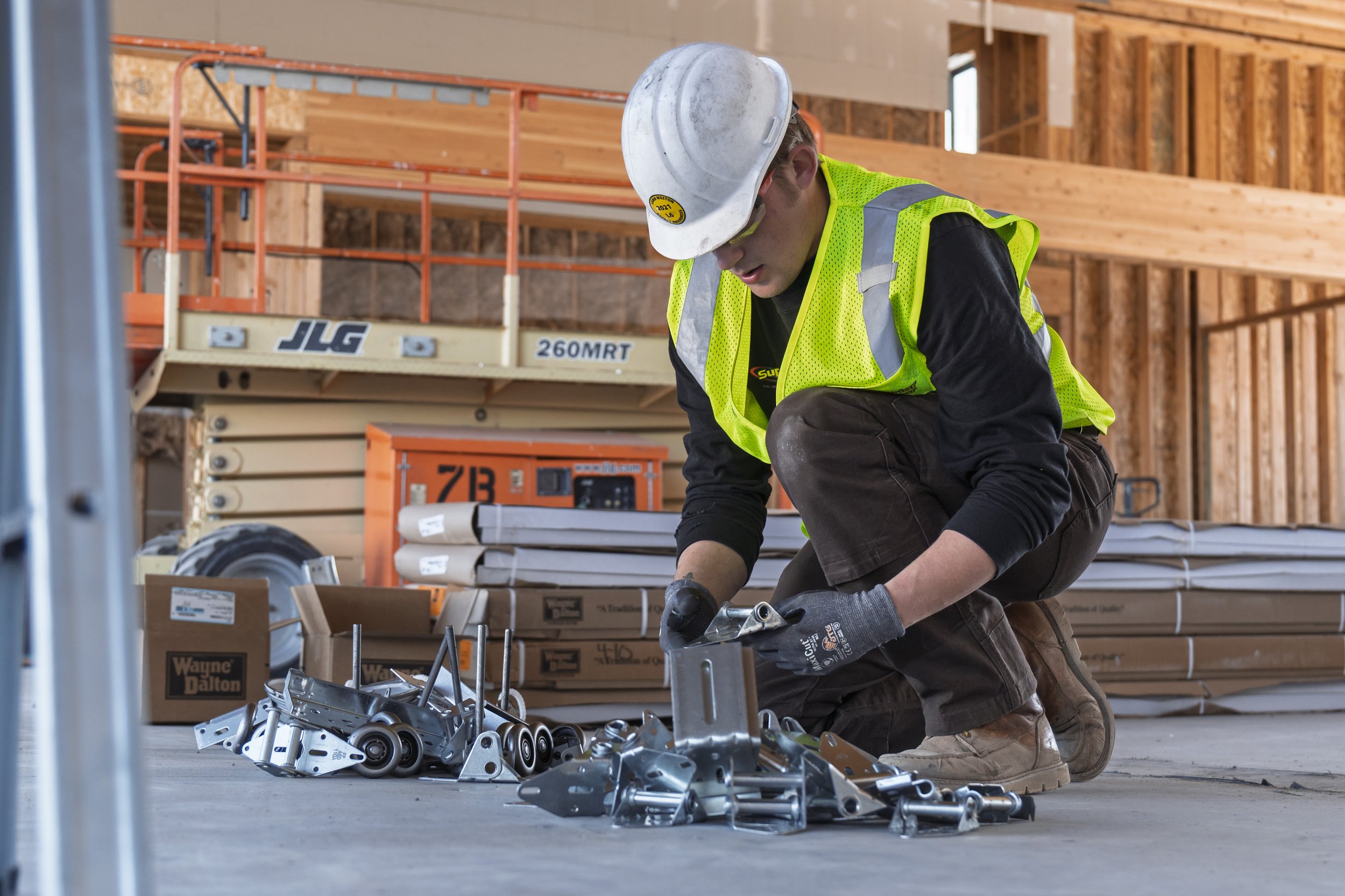 A construction worker in a neon yellow safety vest and a white hard hat kneels on the floor working on metal parts surrounded by boxes and tools in a building under construction.