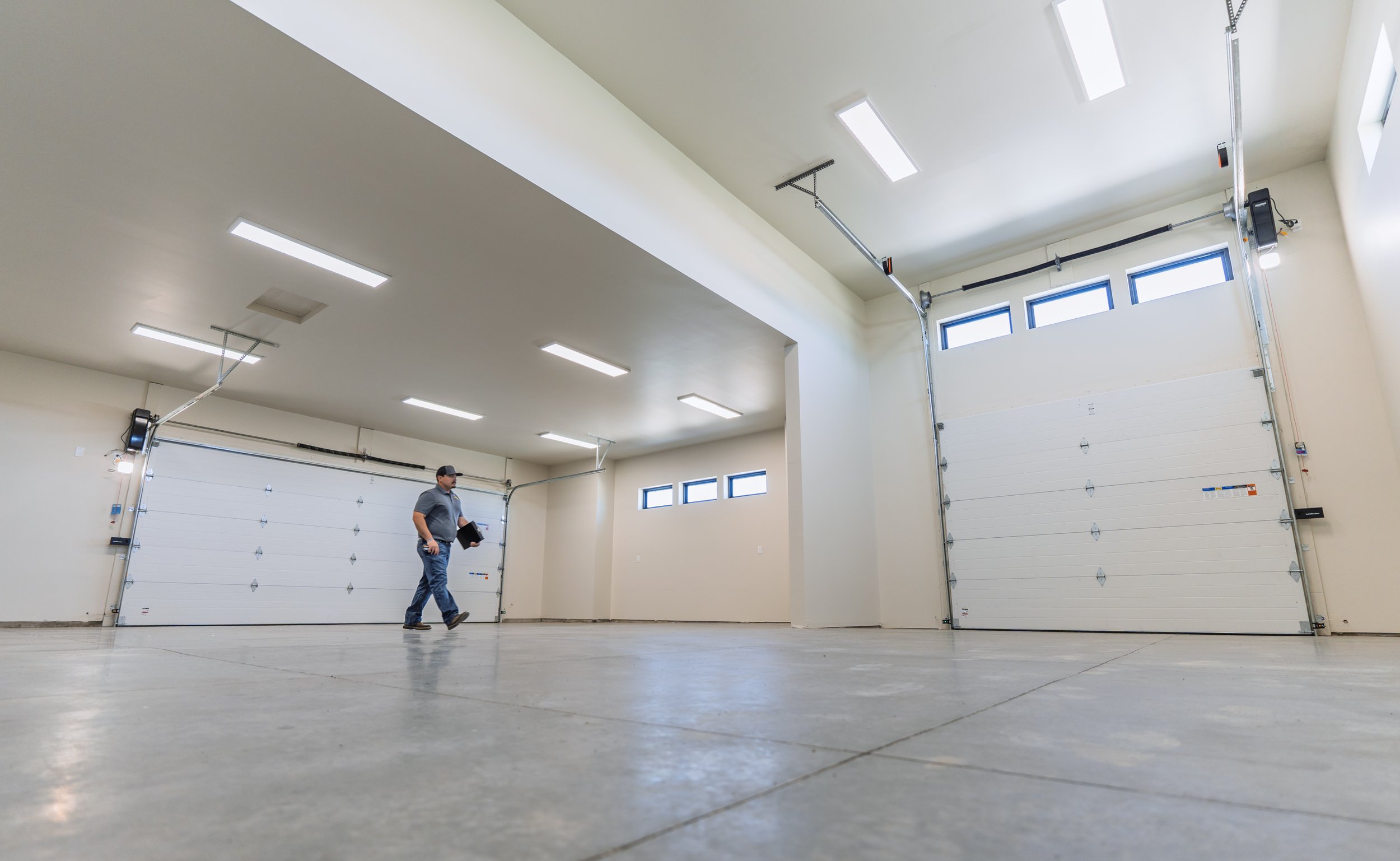 A spacious garage with white walls, a concrete floor, and white overhead garage doors. Fluorescent ceiling lights illuminate the area. A person holding a laptop walks across the open space, and garage door openers are mounted on the ceiling.