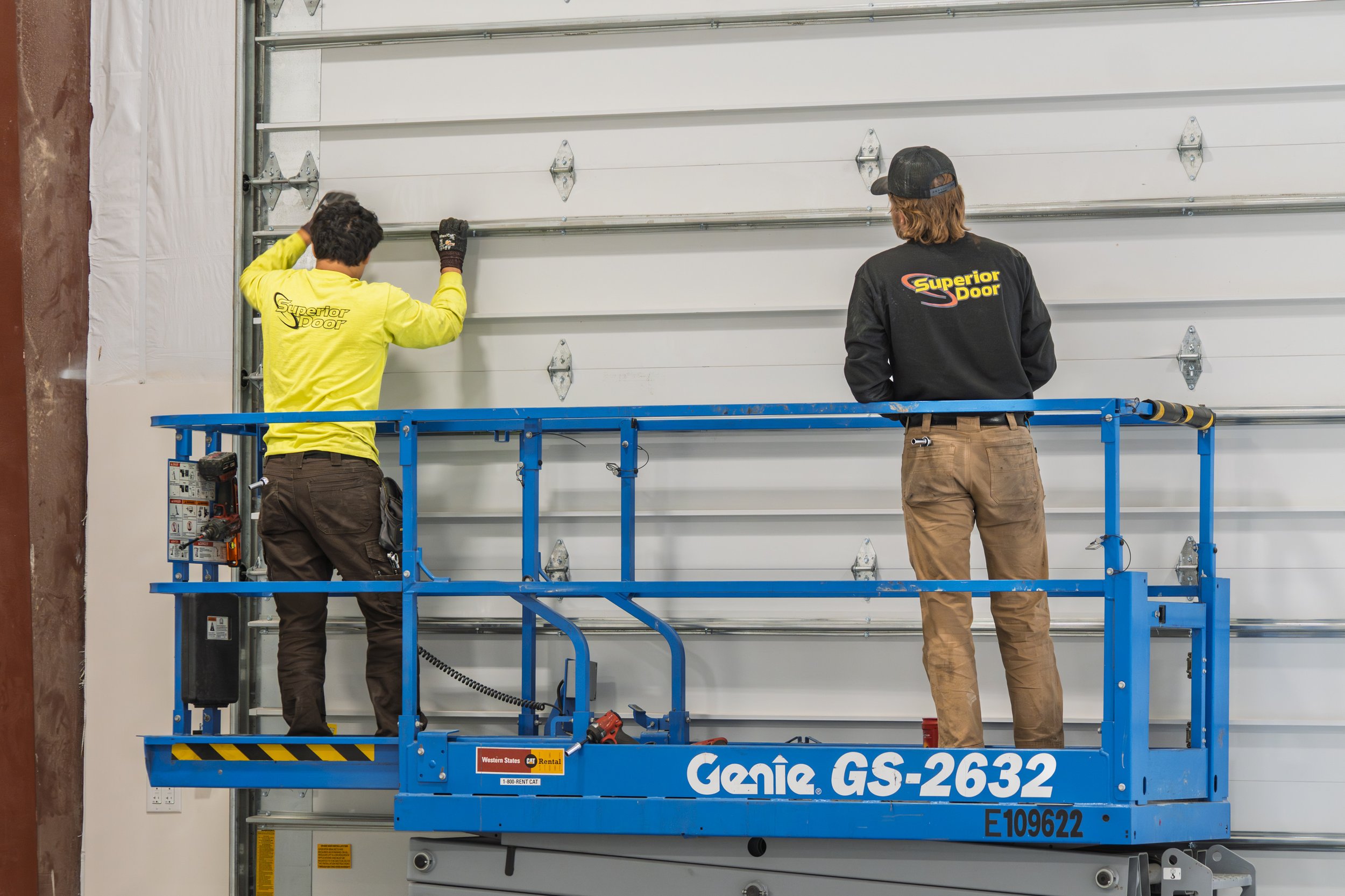 Two workers are installing or repairing a large white garage door using a blue Genie GS-2632 scissor lift. One worker is wearing a yellow shirt and the other a black shirt with the Superior Door logo.