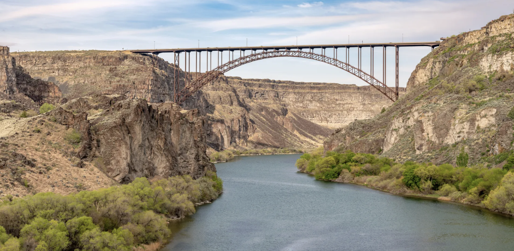The image shows a river flowing through a canyon with steep rocky cliffs on either side. A large steel arch bridge spans the canyon, connecting the two sides. The sky is partly cloudy.