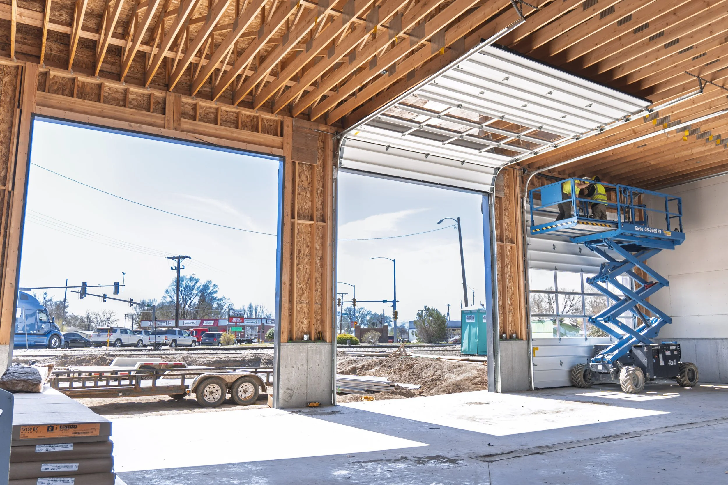 Construction of a building with wooden framing and a partially installed garage door. A worker operates a blue scissor lift near the ceiling. Outside, there is a parking lot with cars and a street with traffic lights.