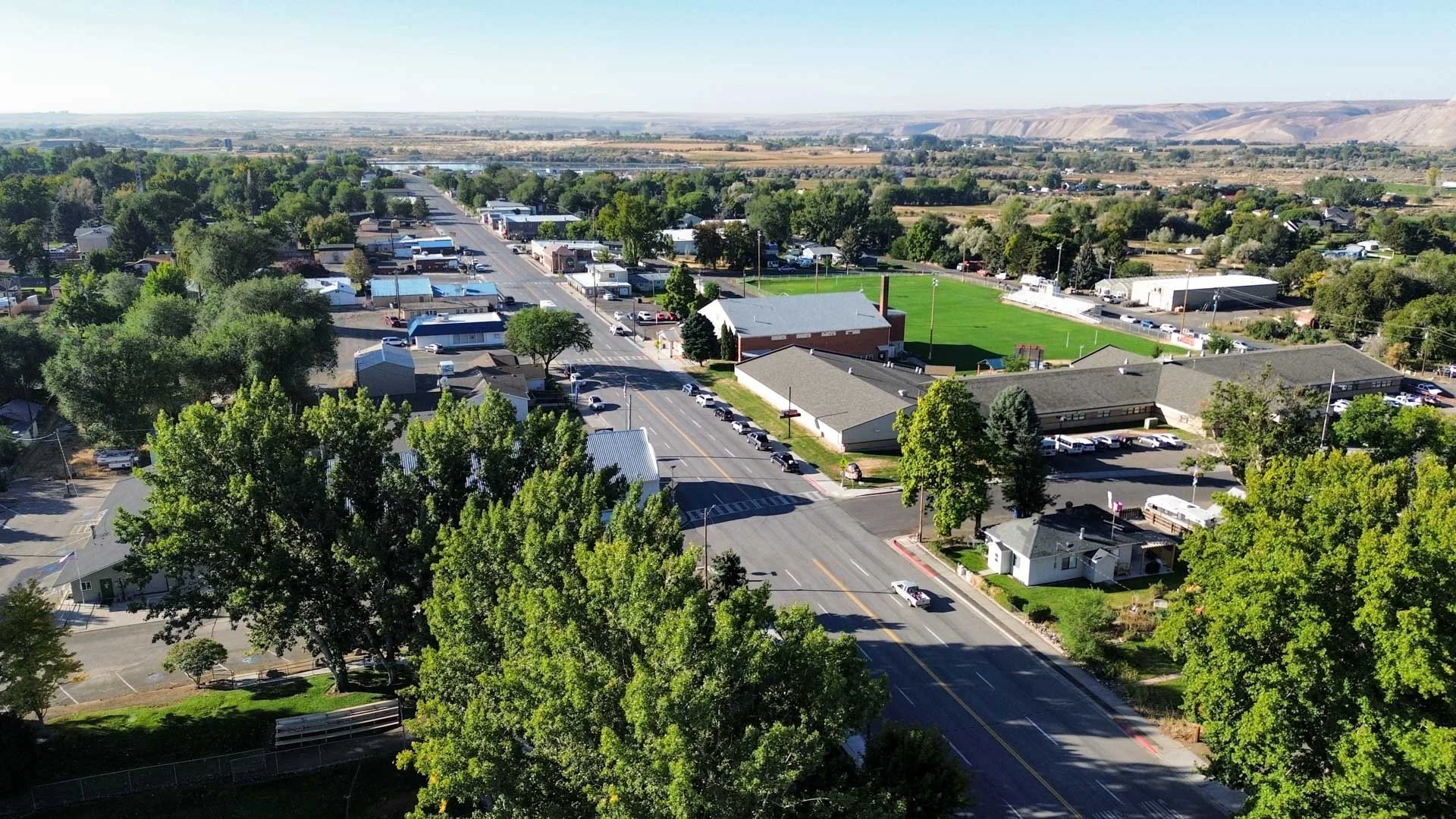An aerial view of a small town with a main street, buildings, green trees, a school with a football field, and surrounding rural landscape with hills in the background.