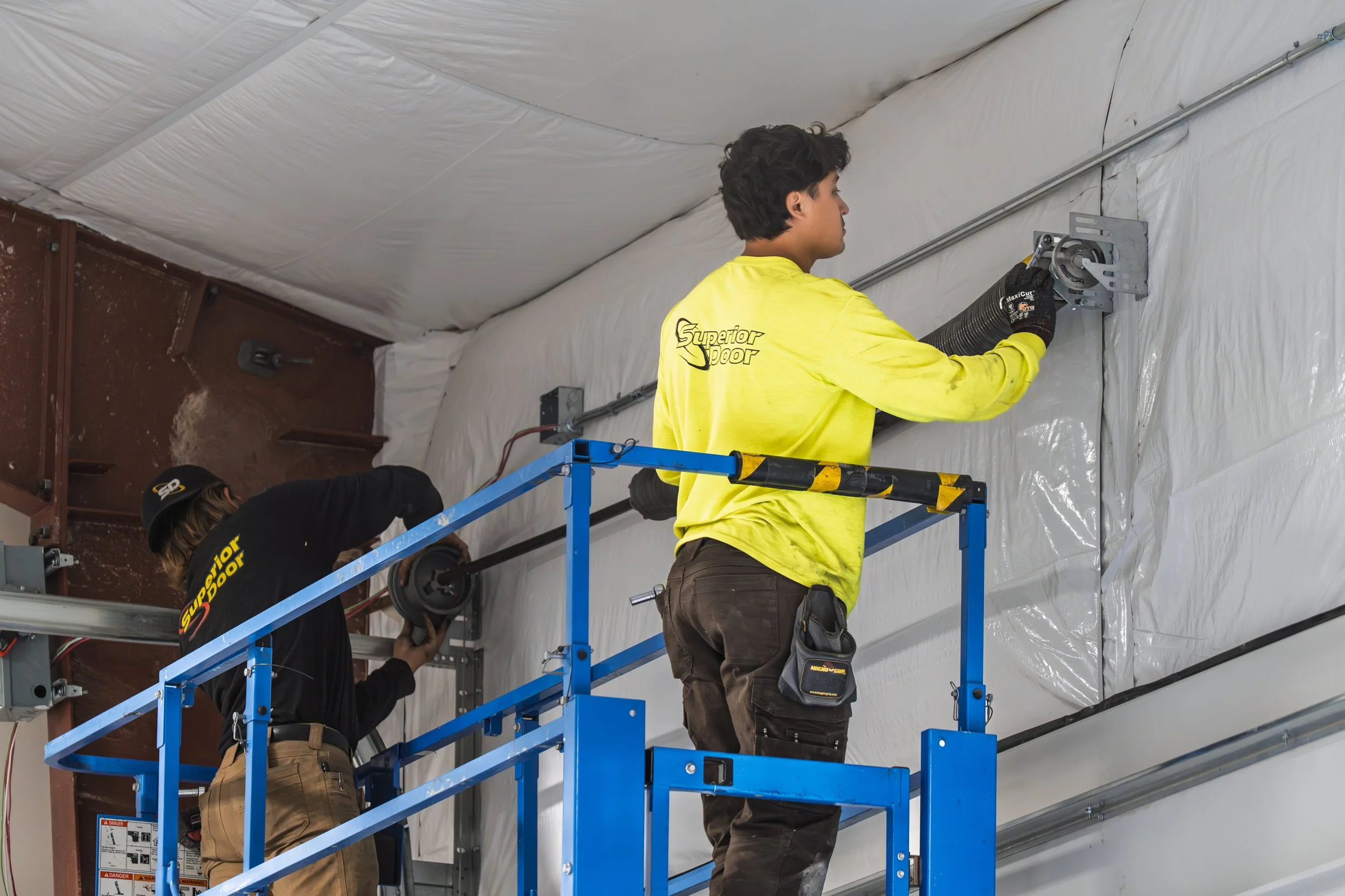 Two workers on a blue lift installing or maintaining a large duct in an industrial or commercial building. They are wearing yellow shirts with "Superior Door" logo and safety gloves, with the ceiling covered in white insulation material.