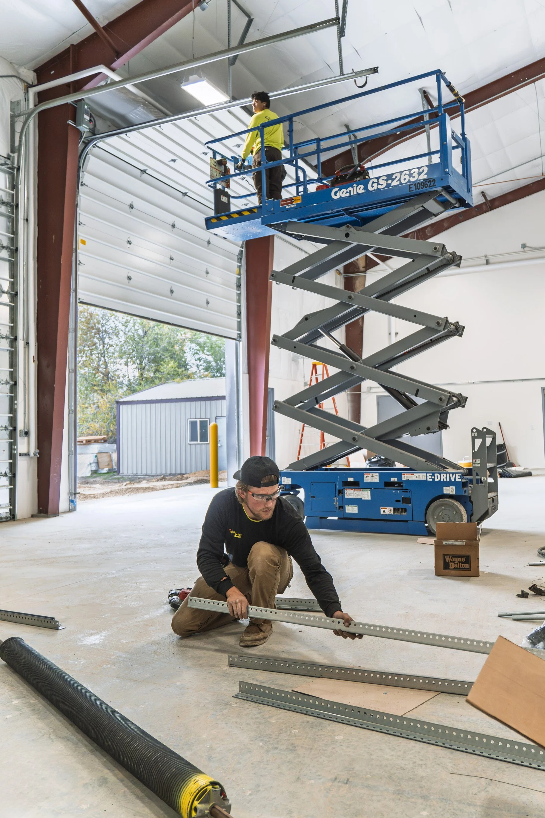 Two workers are installing or assembling metal framing inside a large industrial garage or warehouse with a high ceiling. One worker is kneeling on the floor, measuring or adjusting a long metal piece, while the other is on a blue scissor lift working near the ceiling. Various tools, a box, and electrical wiring are visible in the workspace.