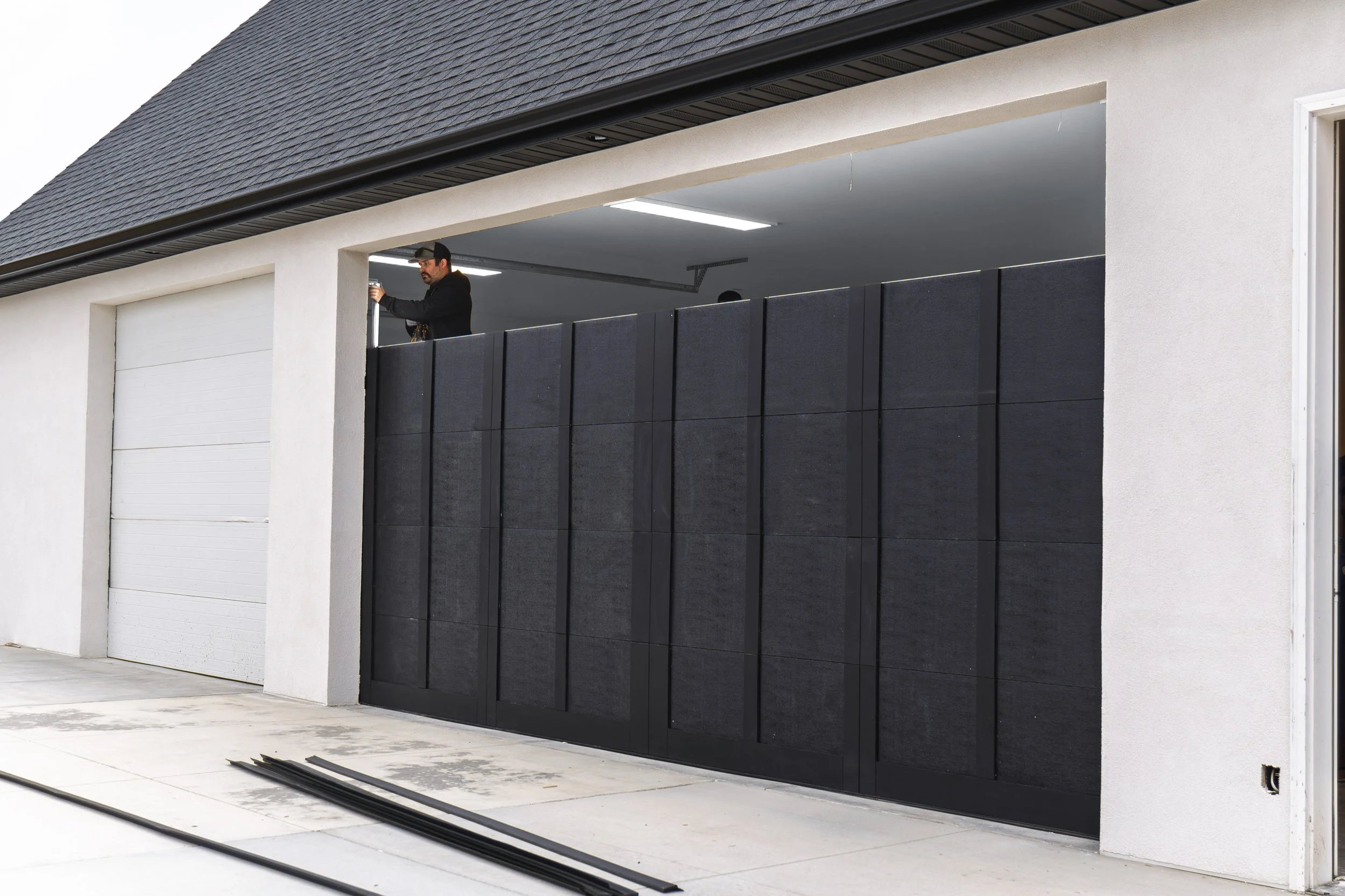 A man installing a black sliding gate in front of a white garage door with a pitched shingled roof.