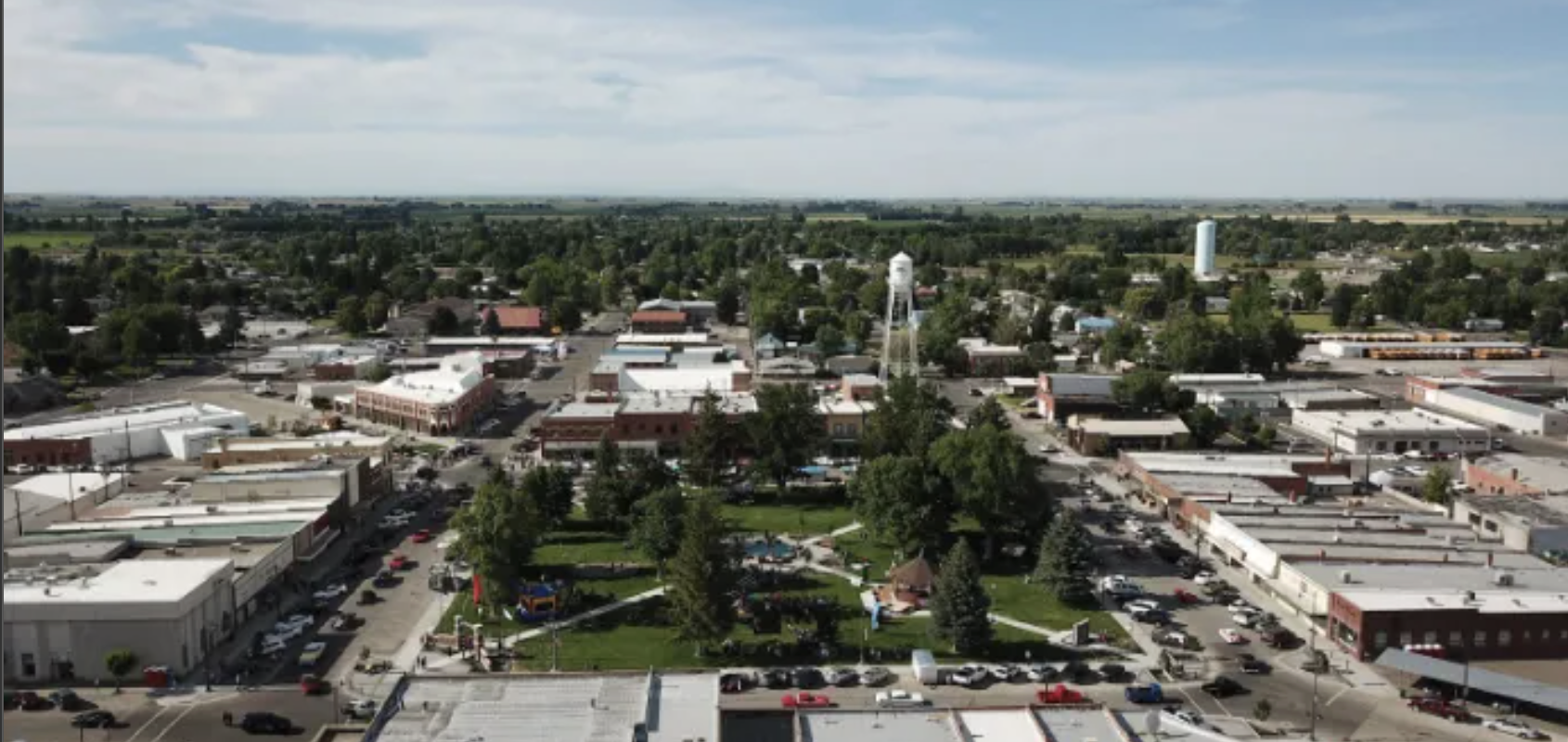 Aerial view of a small town with a central park and surrounding commercial buildings, parking lots, water tower, and a water tower in the distance under partly cloudy skies.