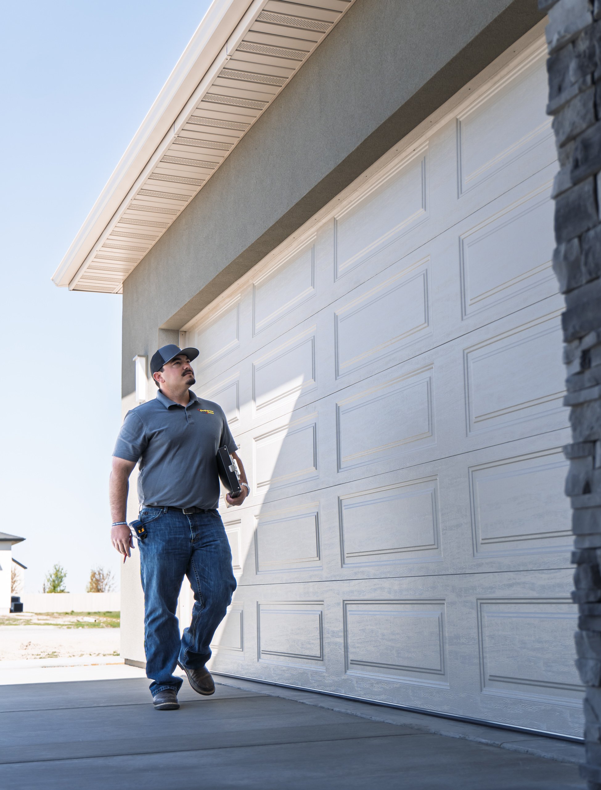 A man wearing a cap and a gray polo shirt is walking past a white garage door on a sunny day, holding a clipboard and some tools.