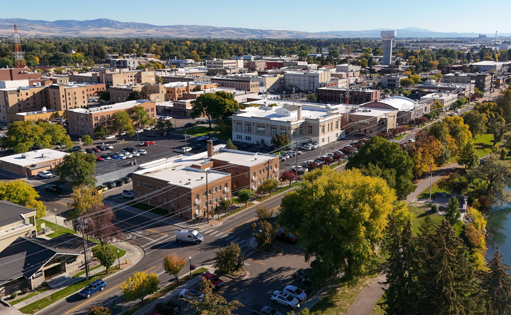 A panoramic view of a city with various buildings, trees with fall foliage, parking lots, and streets, with mountains in the background and a clear blue sky.