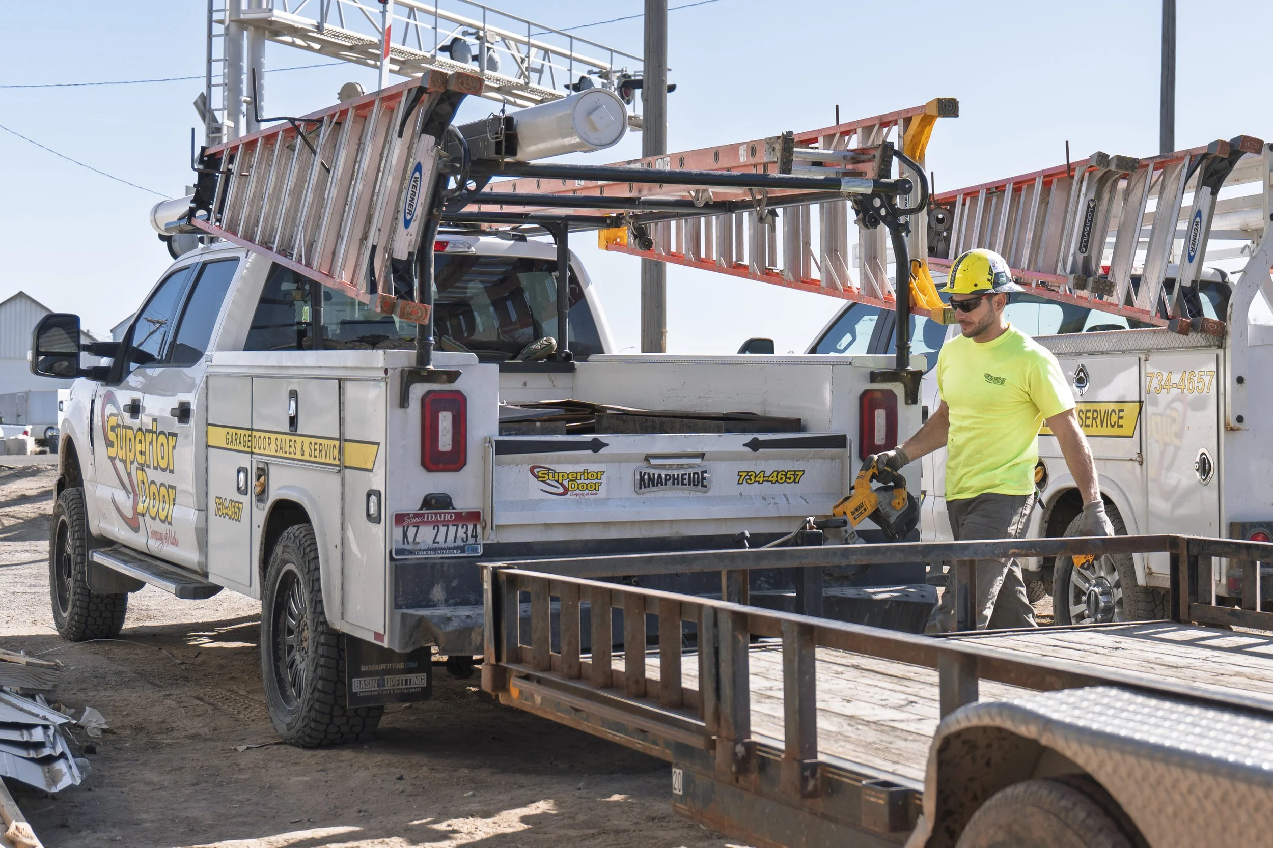 A construction worker wearing a yellow hard hat and sunglasses is walking past a work truck with ladders on top. The truck has Garage Door Sales & Service branding, and the scene appears to be at a construction site with tools and materials around.