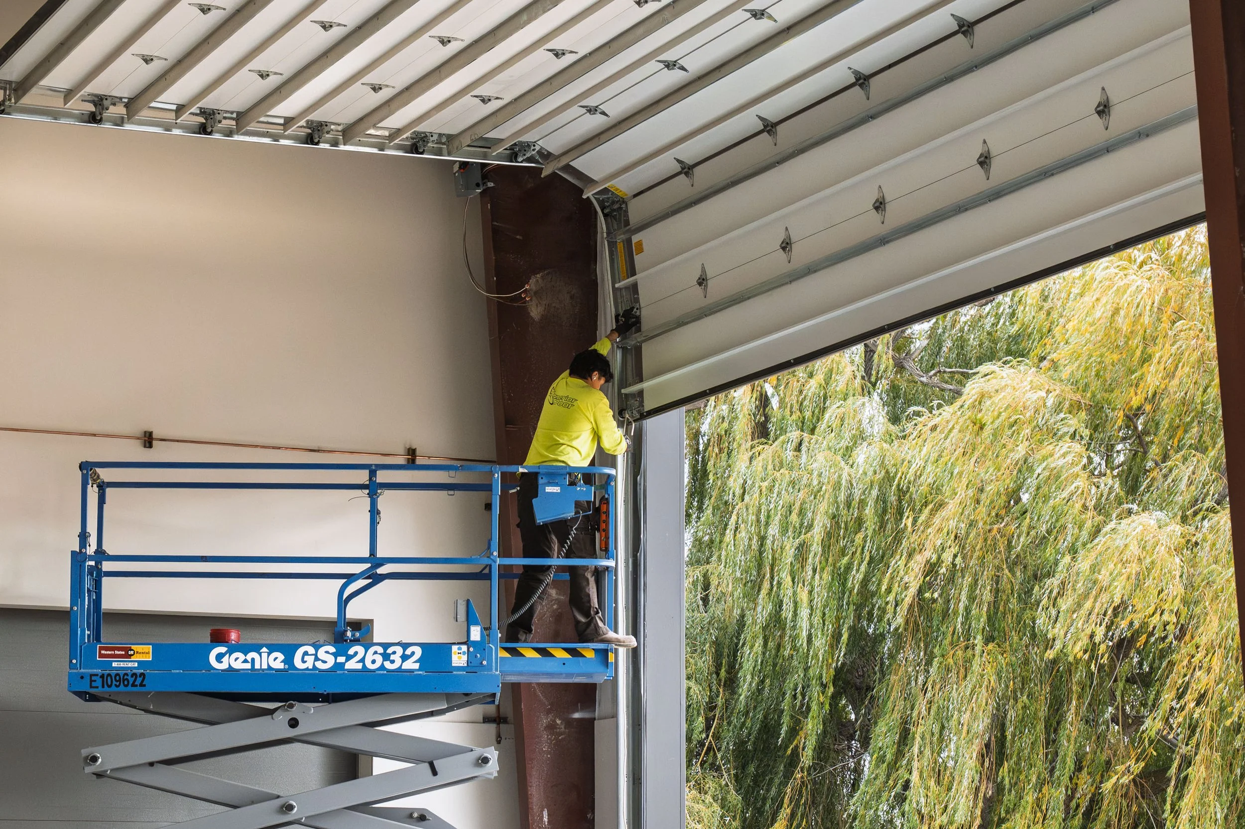 A worker in a yellow shirt on a blue scissor lift inspecting or working on a large, partially installed garage door with a background of green and yellow trees outside.