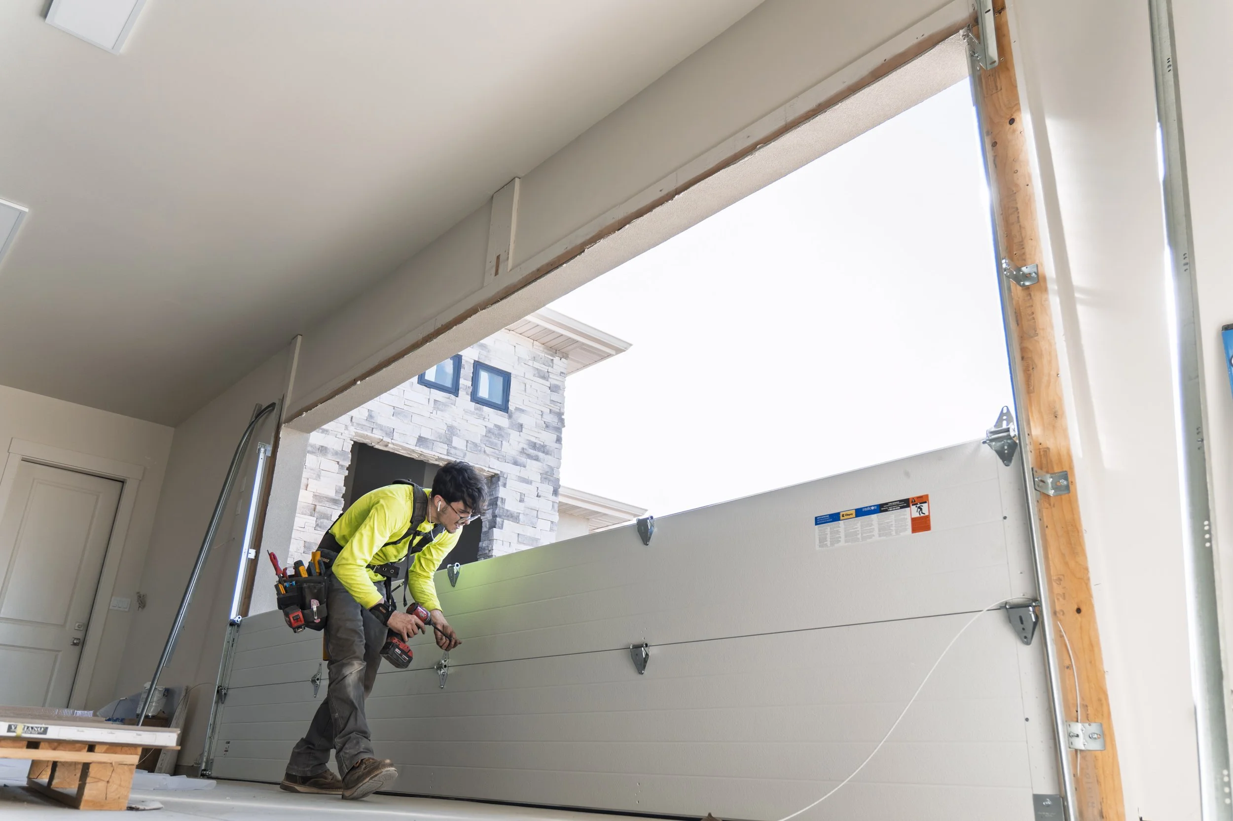 Construction worker installing a garage door with tools on his belt, inside a garage with an open door and visible house exterior.