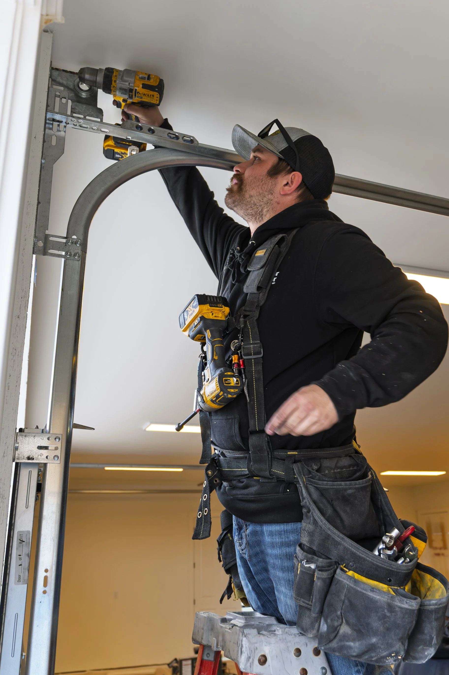 A construction worker is standing on a platform, using a cordless drill to work on a ceiling. The worker is wearing a black hoodie, a tool belt, and a cap with sunglasses on it. The space has bright ceiling lights and plain walls.