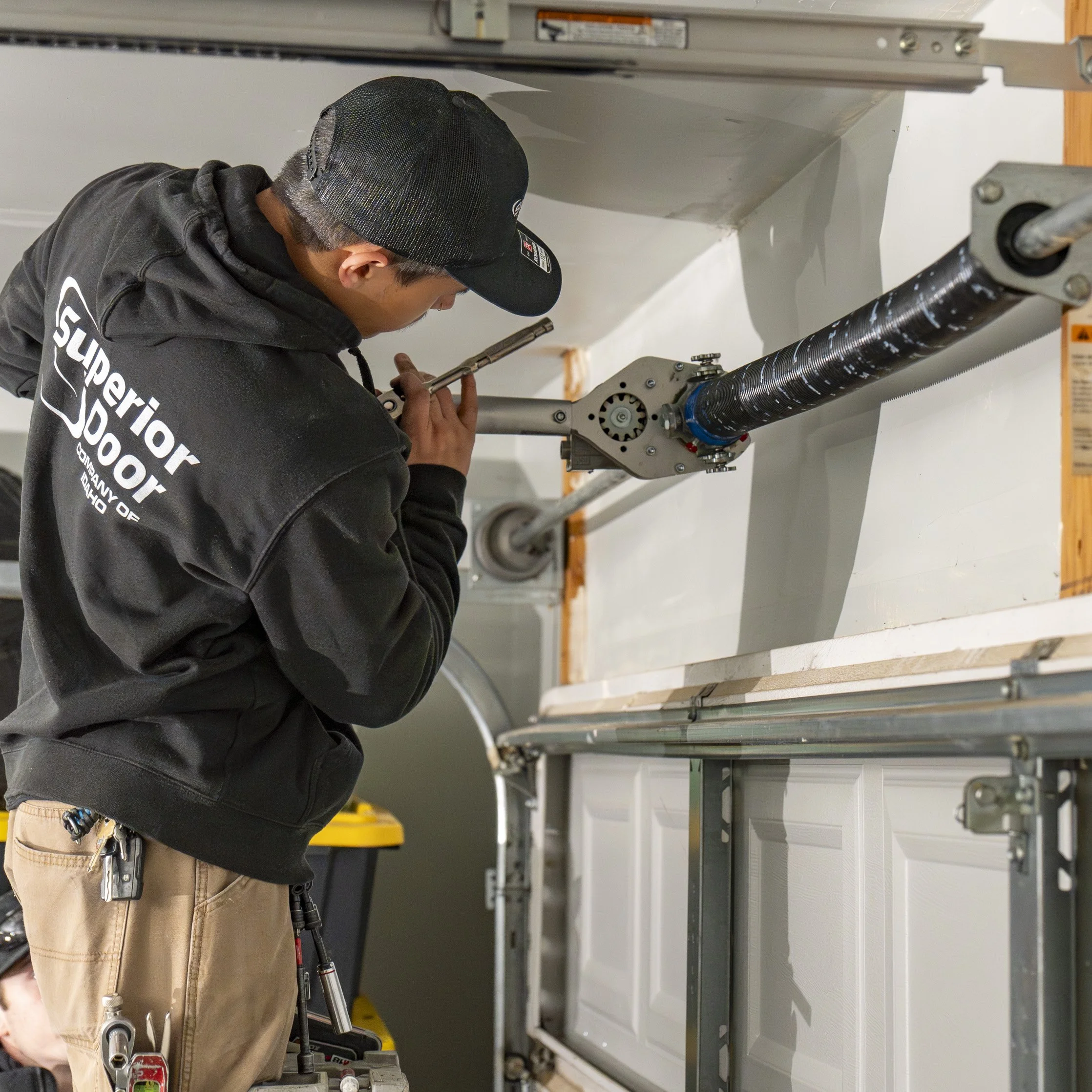 A person wearing a black hoodie, beige pants, and a black mesh cap is working on a mechanical or industrial component using a tool. The person appears to be focused on the task, and there are various mechanical parts and a scissor lift in the background.
