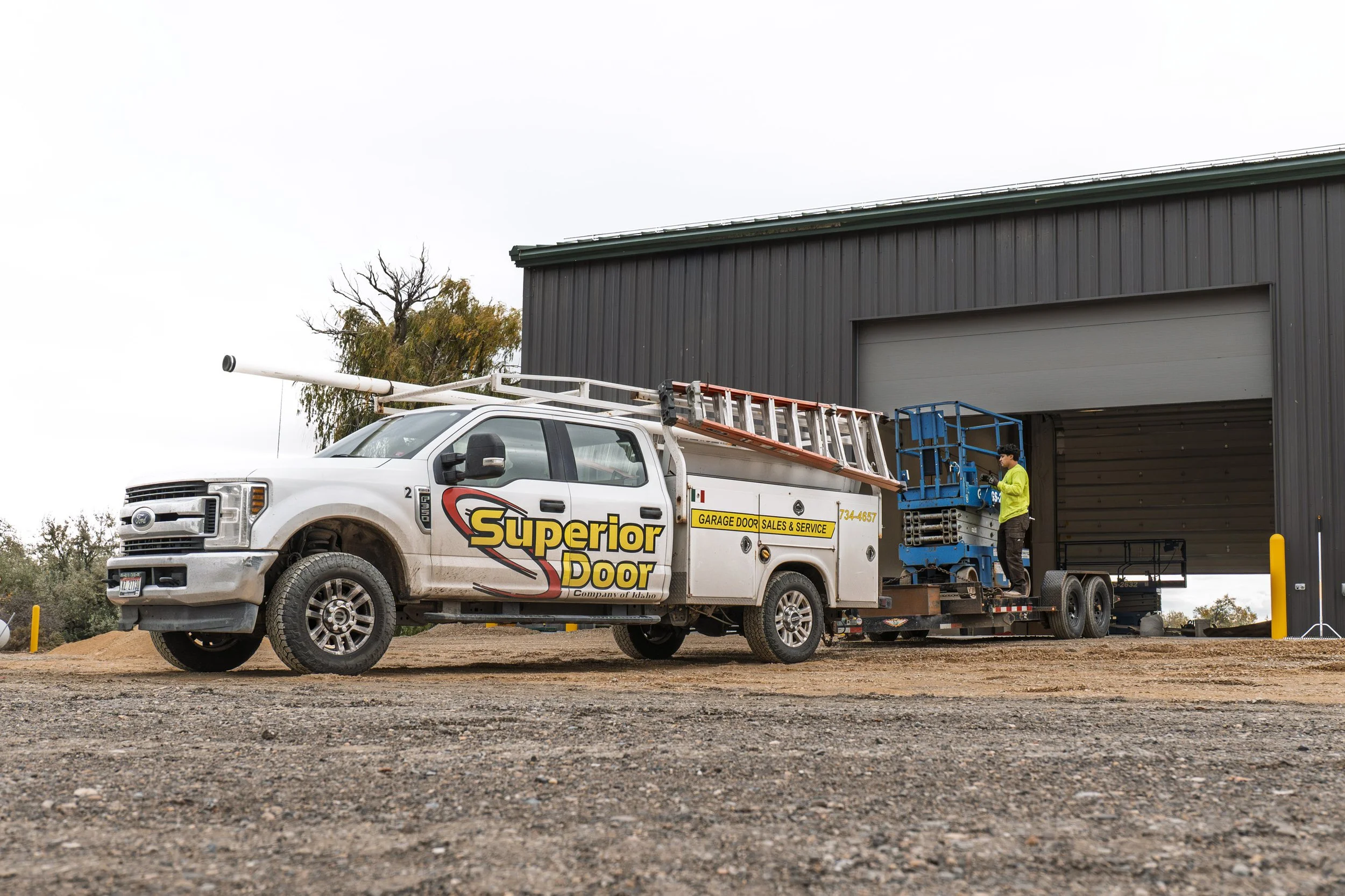 A worker in a safety vest stands next to a service truck parked outside a large gray industrial building, with equipment and ladder on the truck.