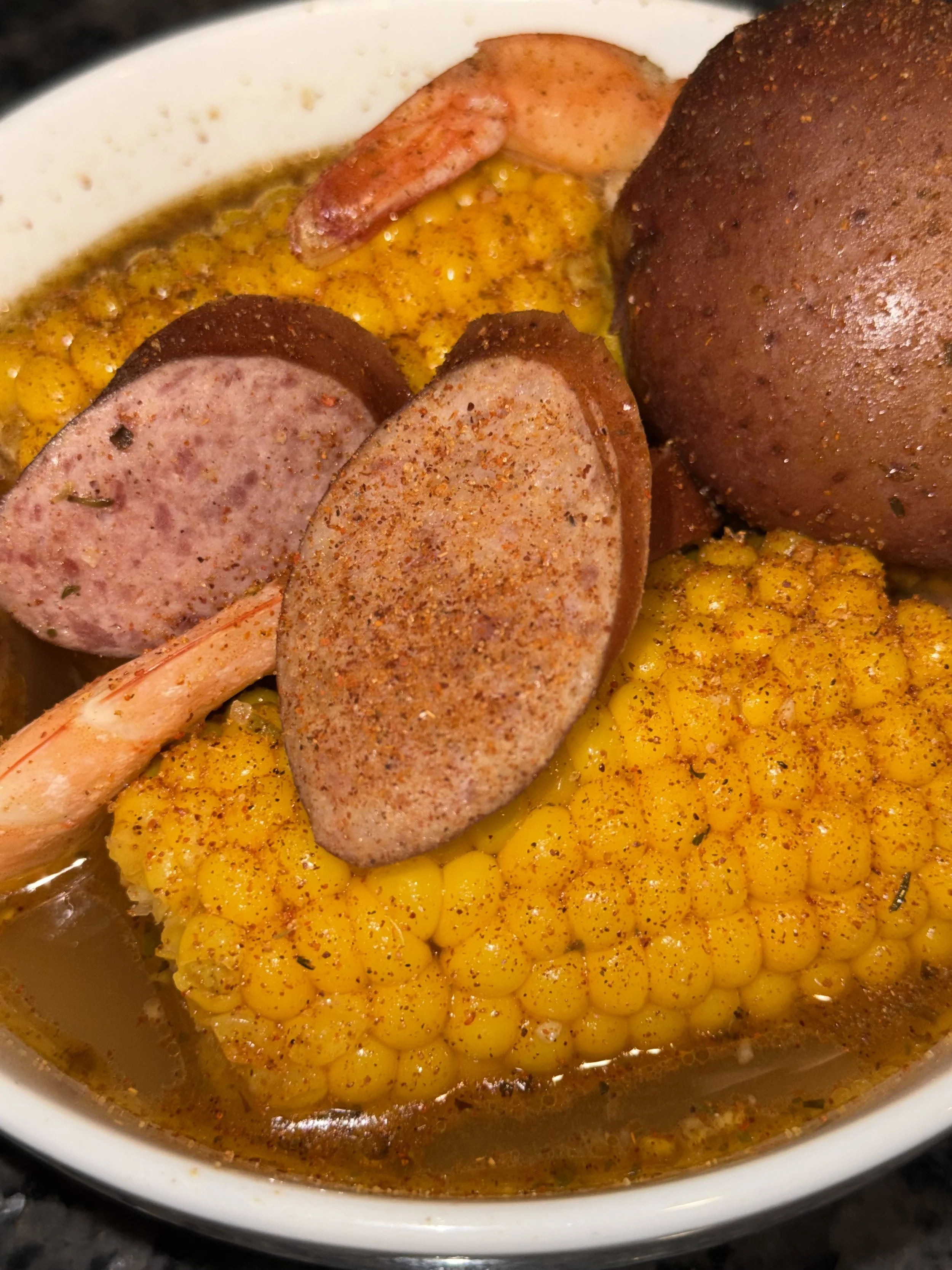 A bowl of cooked corn on the cob garnished with spices, slices of sausage, and a seasoned piece of meat, all submerged in broth or sauce.