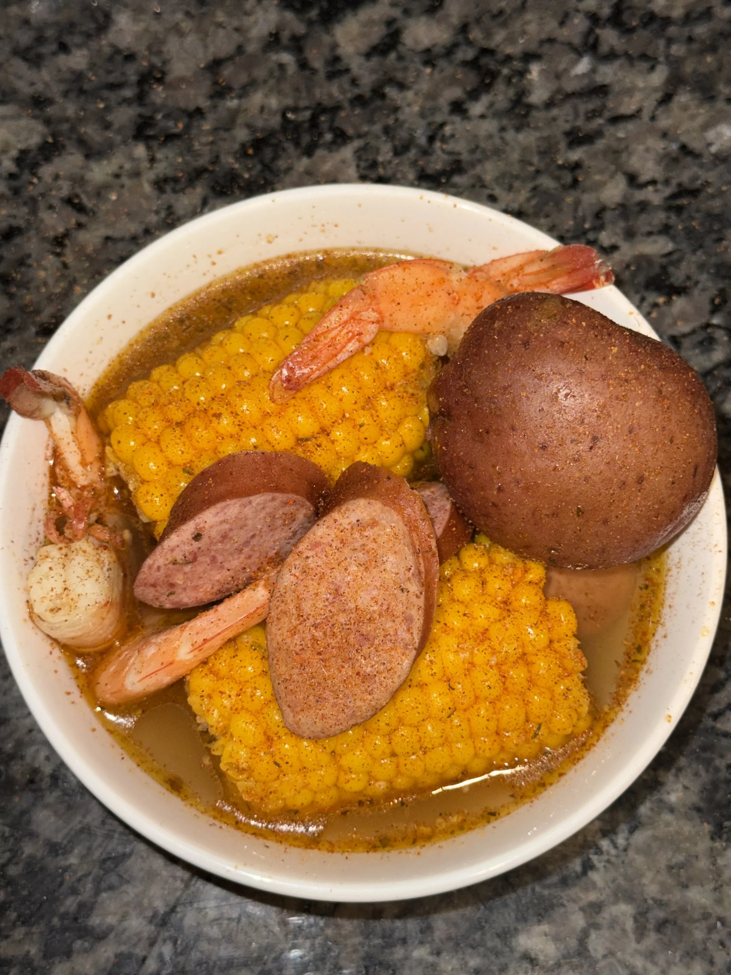 A bowl of chili with yellow corn, shrimp, sliced sausage, and a whole boiled egg, all on a dark granite countertop.