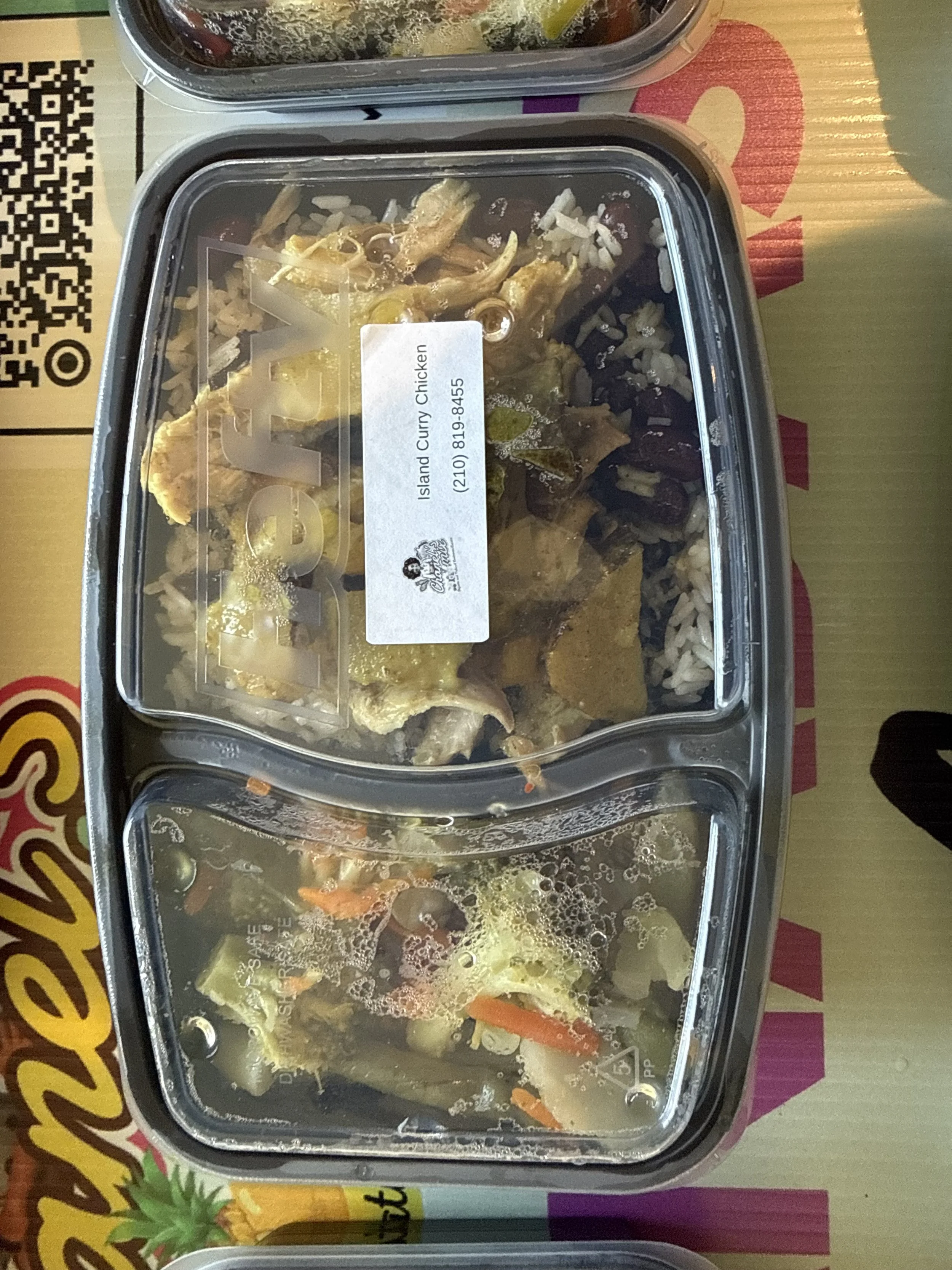 A plastic container of Island Curry Chicken with rice, vegetables, and sauce, placed on a colored cardboard surface.