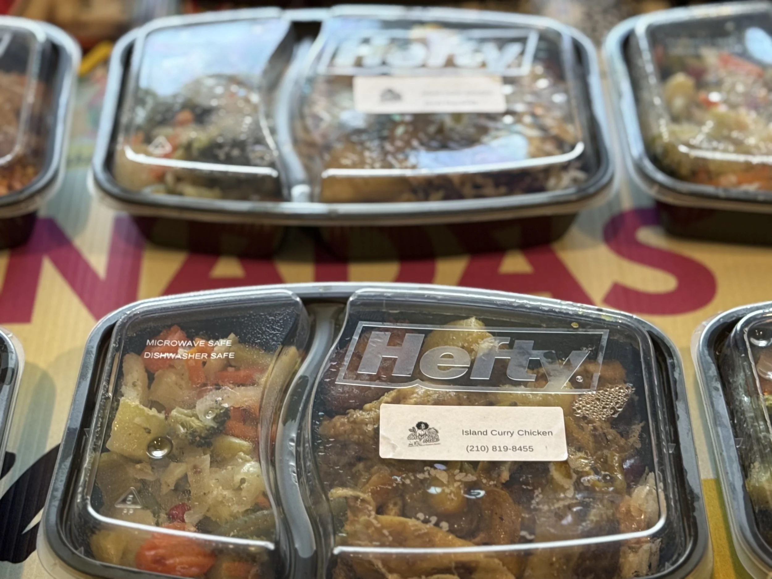A plastic food container labeled 'Island Curry Chicken' with a phone number underneath, placed among other similar containers on a table with a patterned tablecloth.