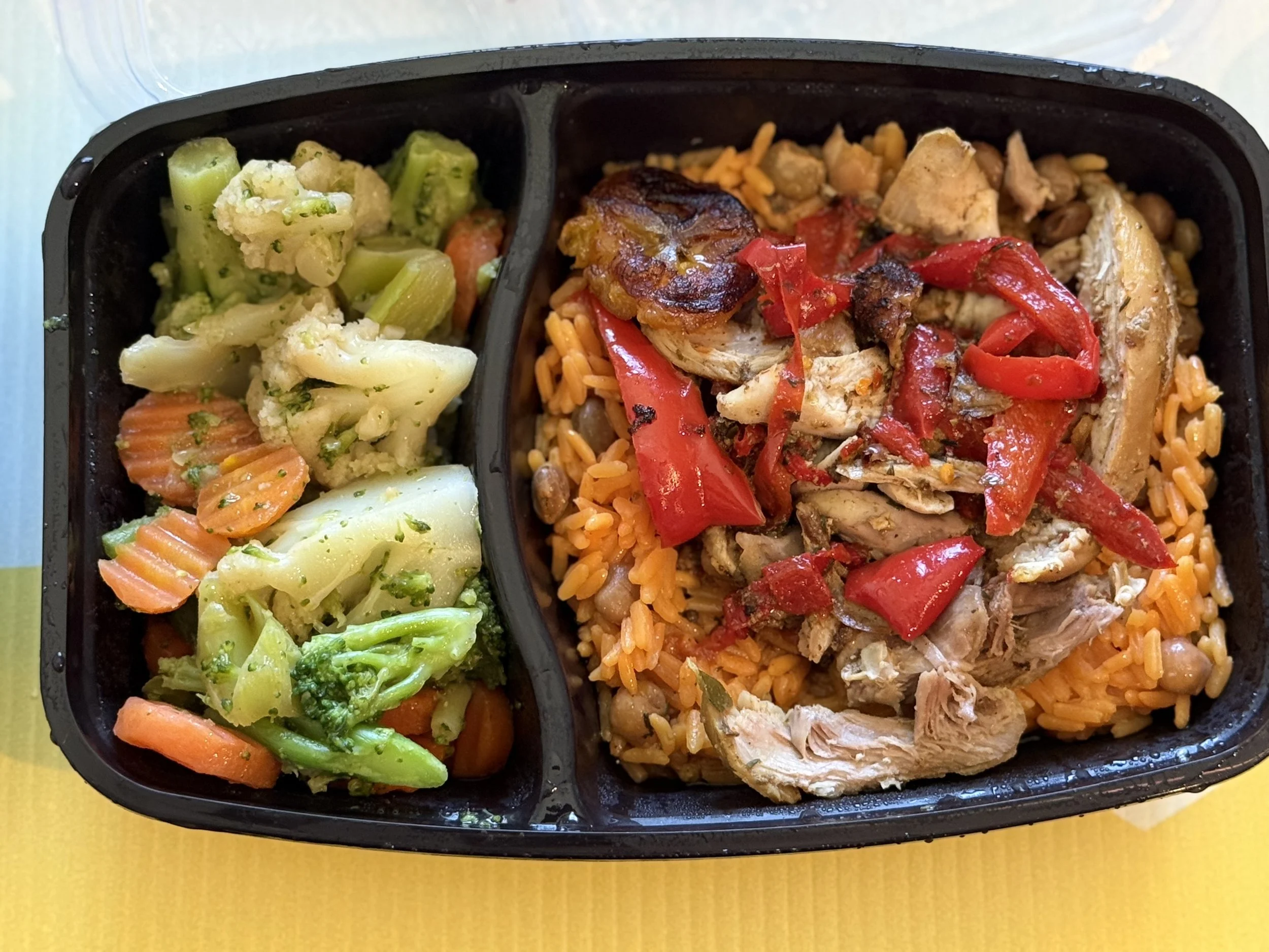 A black compartmentalized meal container filled with vegetables on the left side and rice, chicken, and peppers on the right side, placed on a yellow surface.