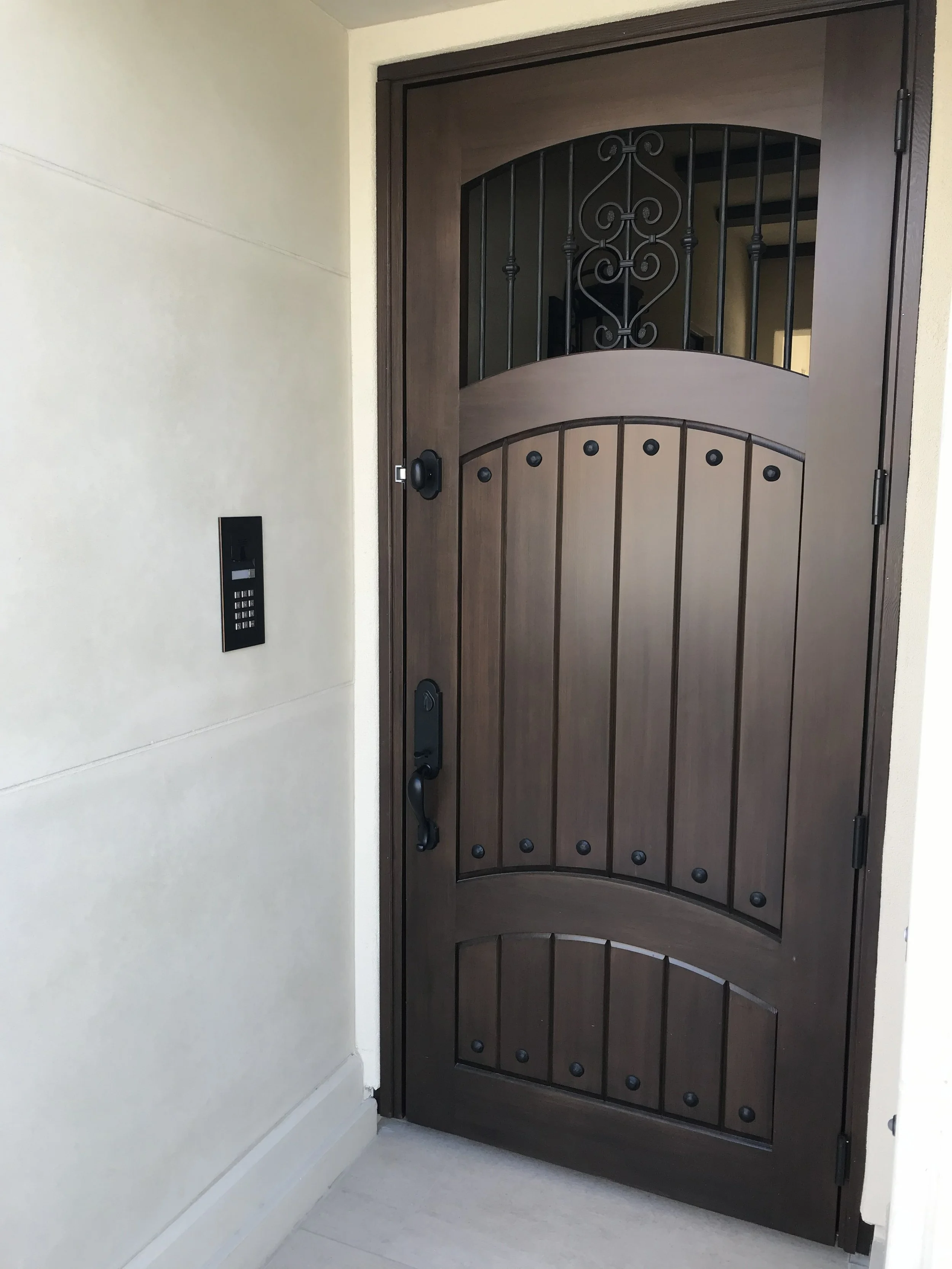 A dark brown wooden front door with decorative ironwork, black handle, and lock, set in a white wall with a black keypad entry system on the left side, and a light-colored tiled floor.