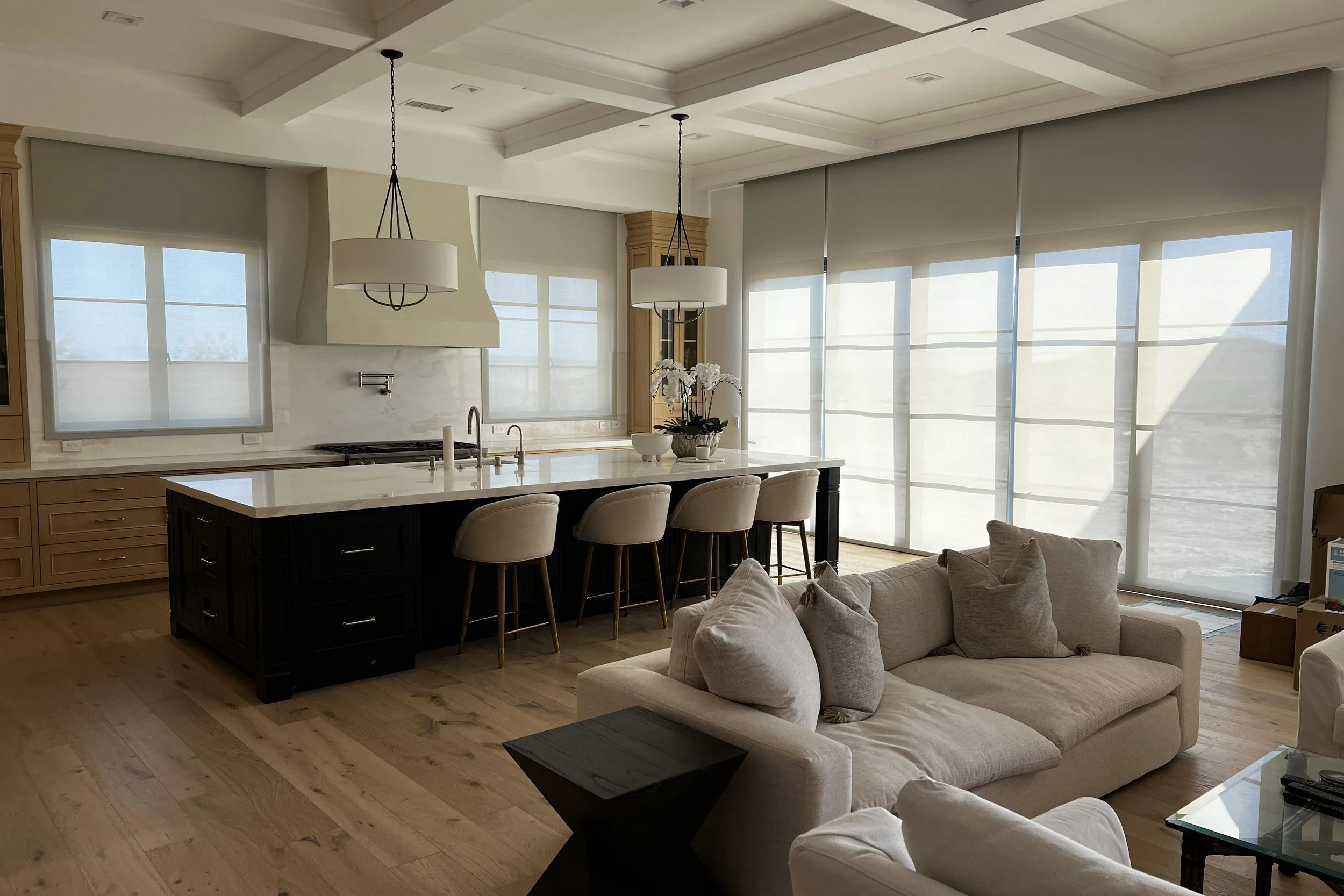 A modern open-concept kitchen and living room with large windows covered by white blinds, a kitchen island with beige upholstered chairs, a beige sofa with plush cushions, and decorative items including a potted orchid on the island and a bowl. Light