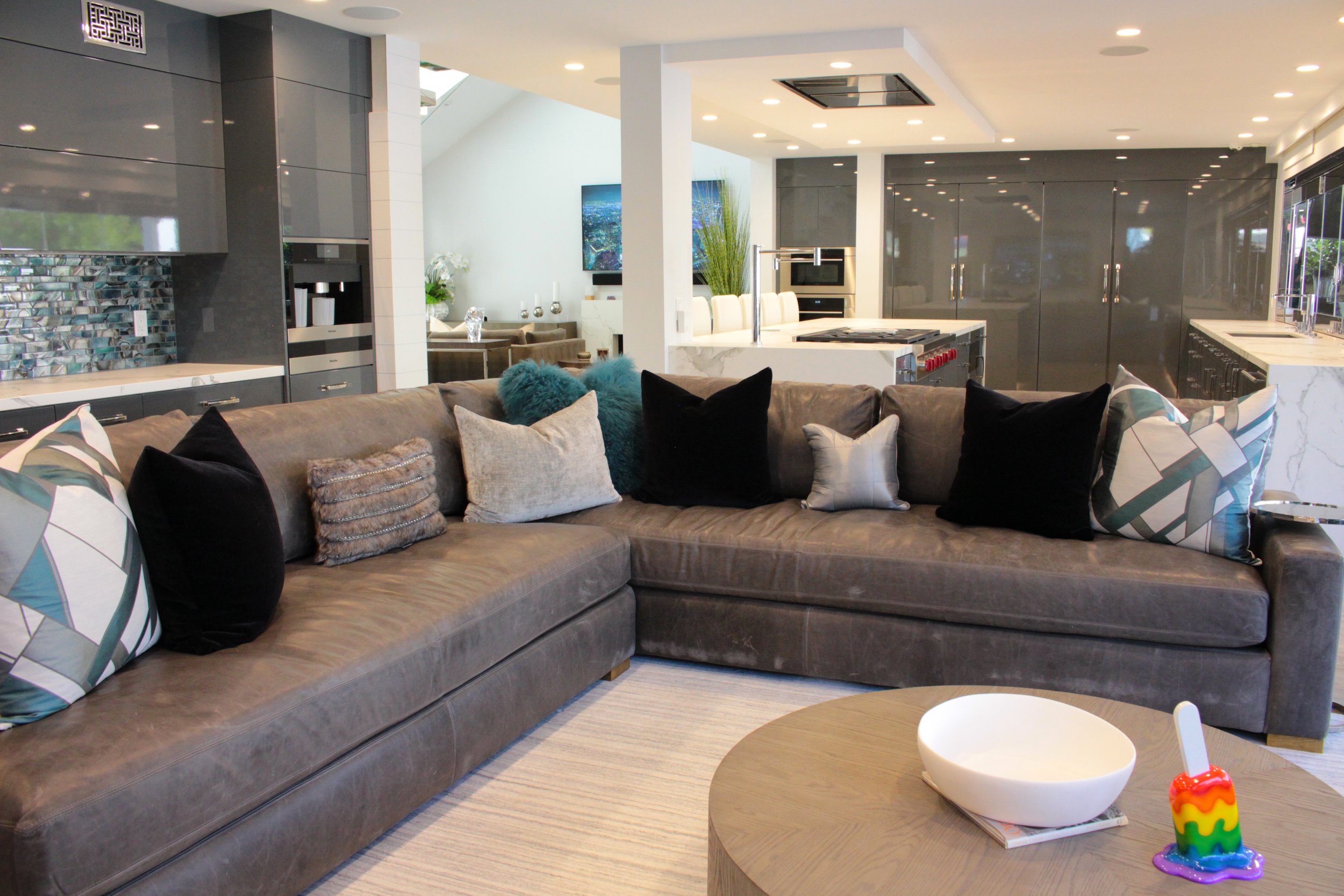 Modern open-concept living room and kitchen with a large gray sectional sofa decorated with decorative pillows, a white marble kitchen island with a built-in stove, sleek gray cabinetry, and a dining area with a table and chairs. The room has bright 