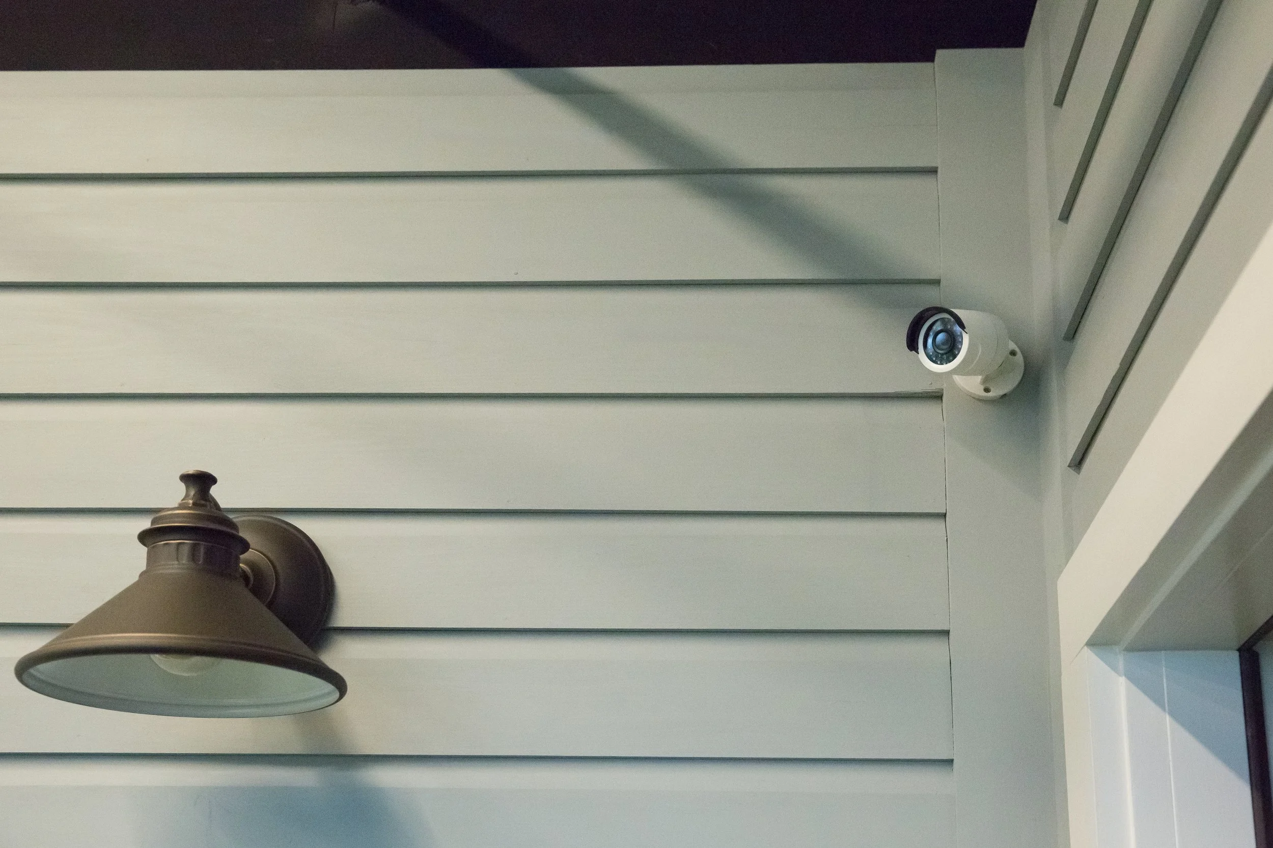A white security camera mounted on the ceiling corner, facing downward. Part of a ceiling fan light fixture with a frosted glass shade and metal details is visible in the lower part of the image.