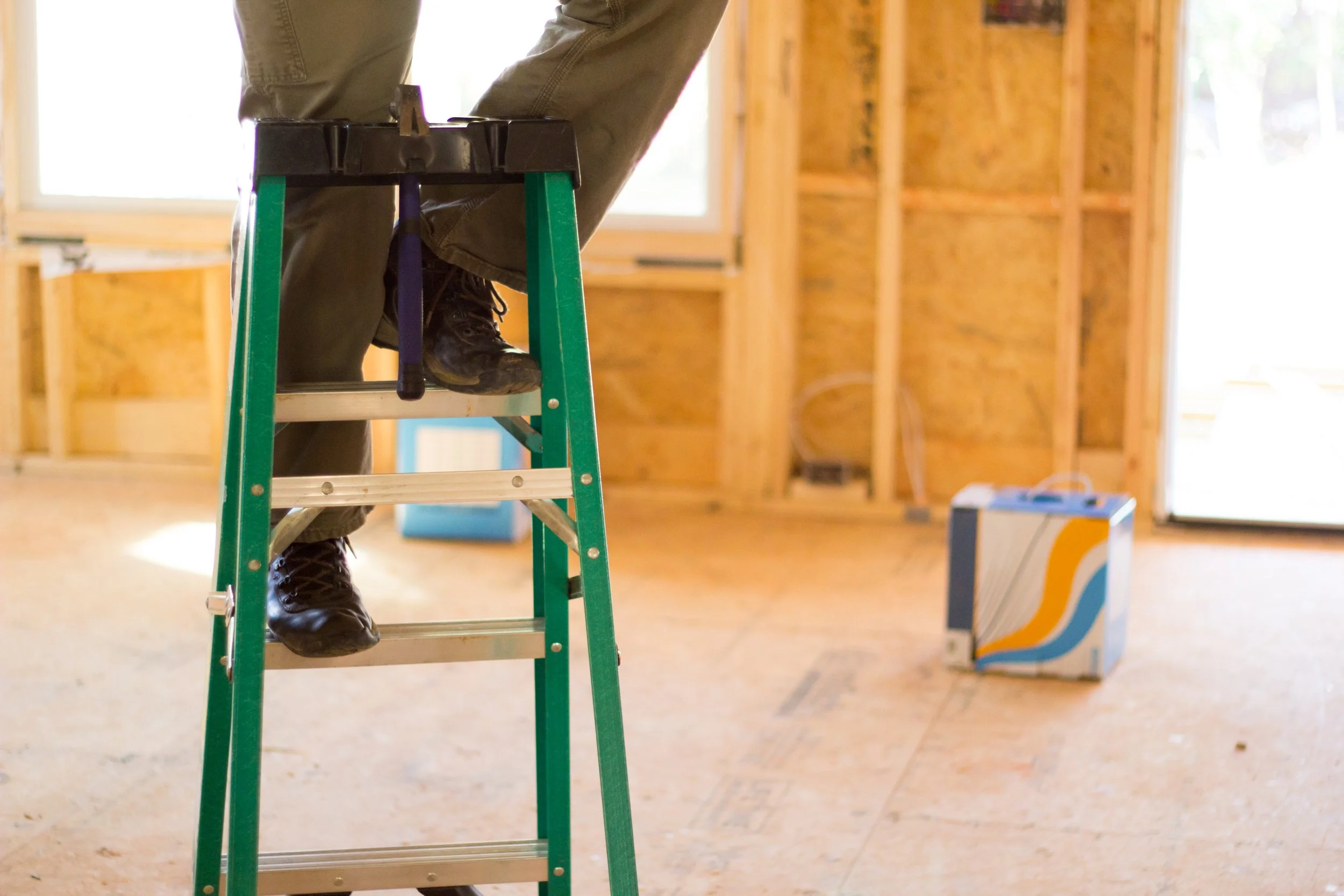 Person standing on a ladder inside a house under construction, with wooden framing and construction materials visible.