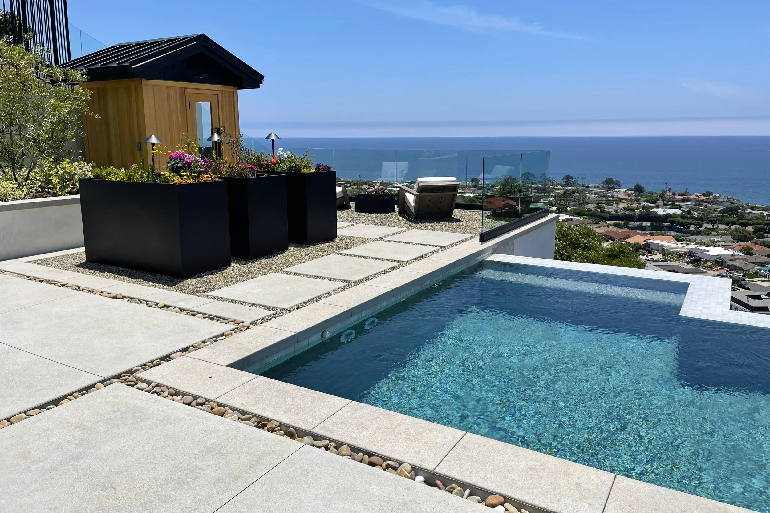 A rooftop patio with a swimming pool overlooking a coastal city and the ocean on a sunny day. The patio has modern outdoor furniture and potted flowers.
